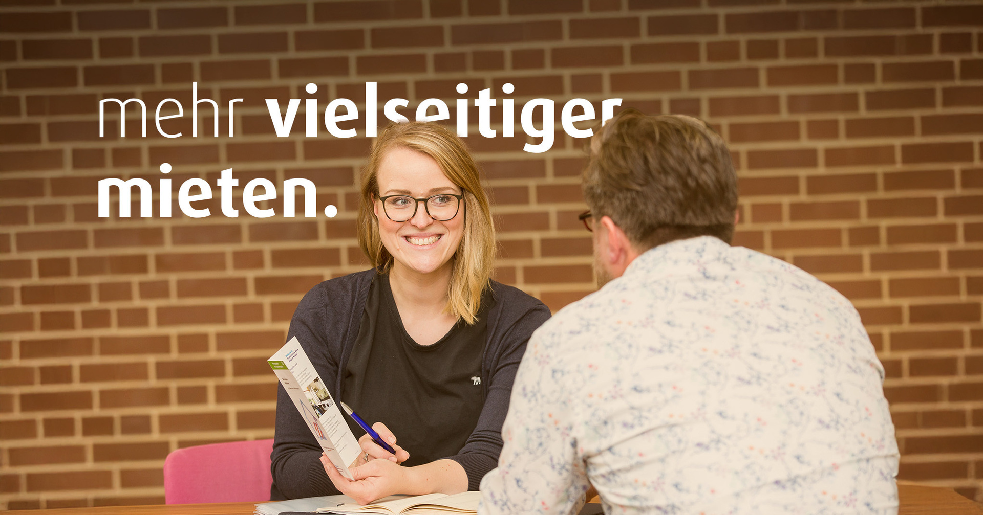 Eine Frau mit Brille und ein Mann sitzen sich gegenüber am Tisch und unterhalten sich. Die Frau zeigt dem Mann eine Broschüre. Auf dem Bild steht der Text "mehr vielseitiger mieten".