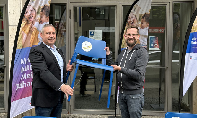Zwei Männer halten einen blauen Stuhl, im Vordergrund zwei weitere blaue Stühle