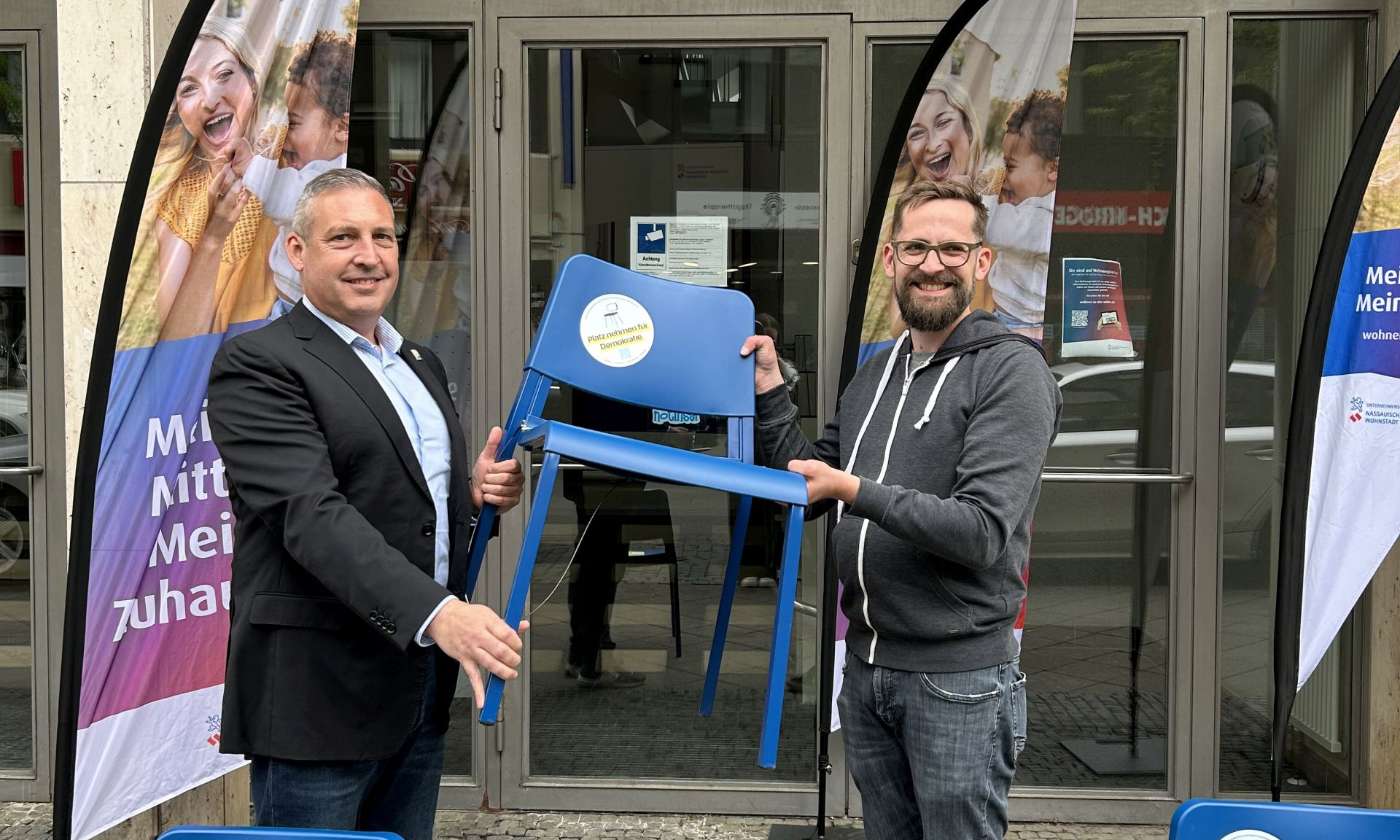 Zwei Männer halten einen blauen Stuhl, im Vordergrund zwei weitere blaue Stühle