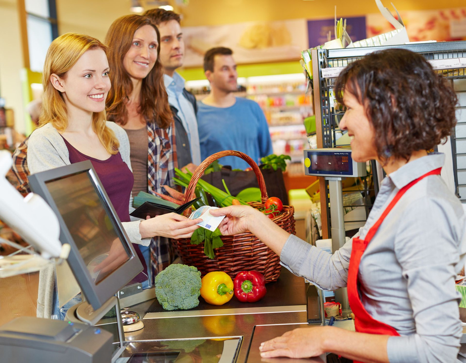 Mehrere Personen stehen an Kasse im Supermarkt. Eine Frau bezahlt in bar.