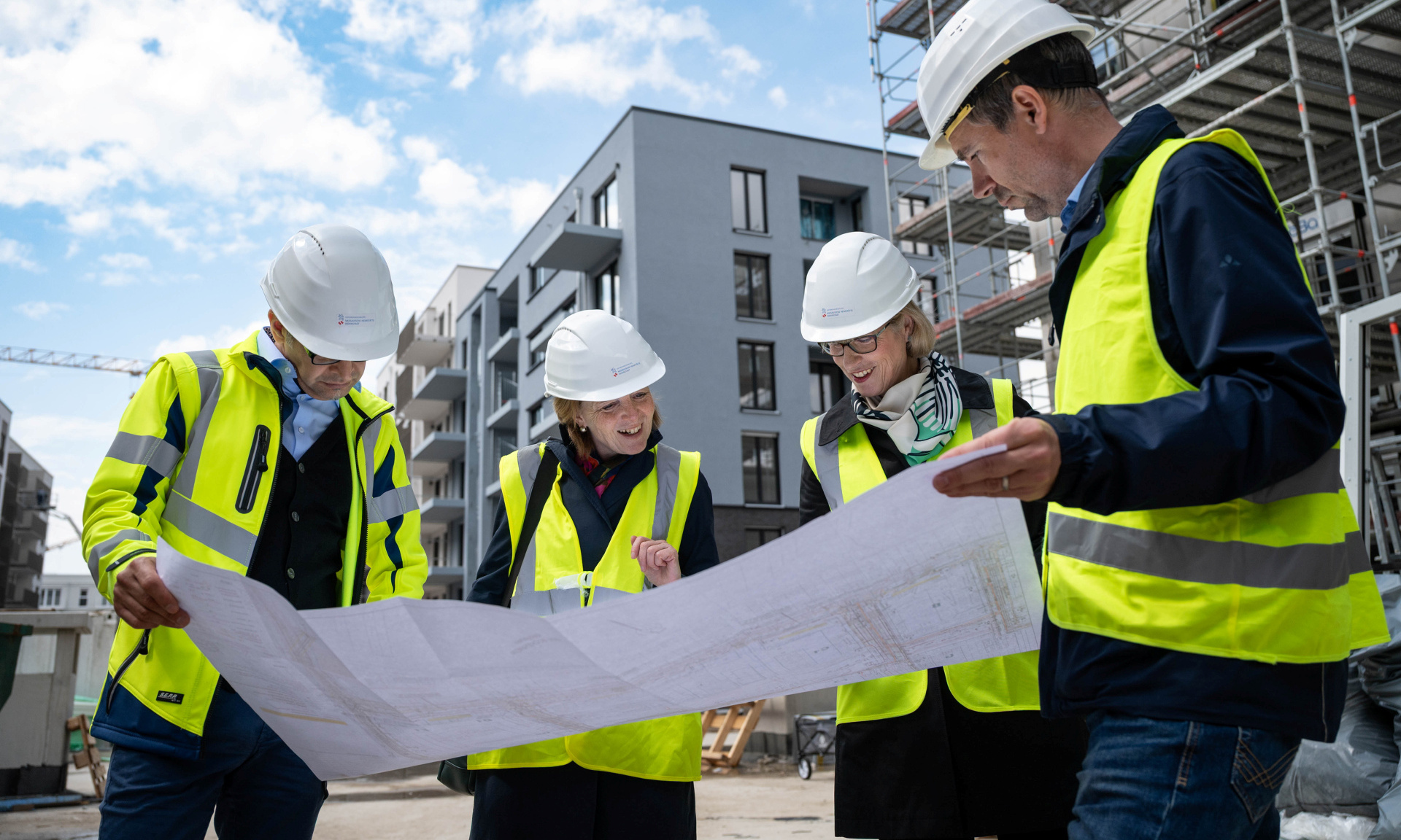 Vier Personen mit Warnweste und weißen Helmen beugen sich über einen großen Bauplan. Im Hintergrund ist Neubau Gebiet zu sehen.