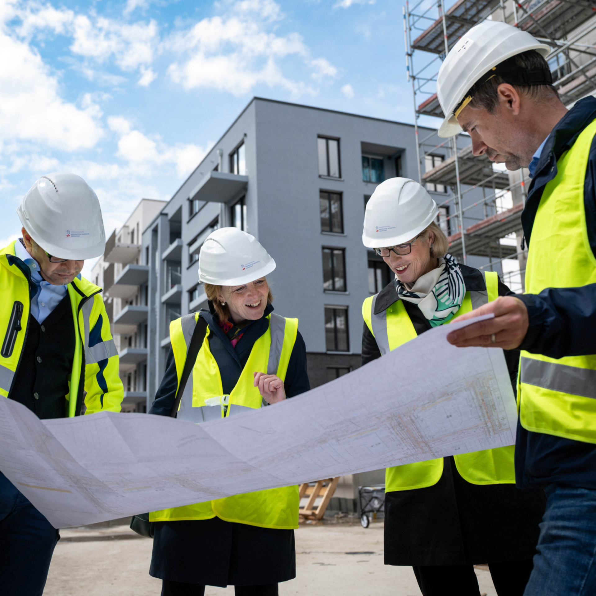 Vier Personen mit Warnweste und weißen Helmen beugen sich über einen großen Bauplan. Im Hintergrund ist Neubau Gebiet zu sehen.