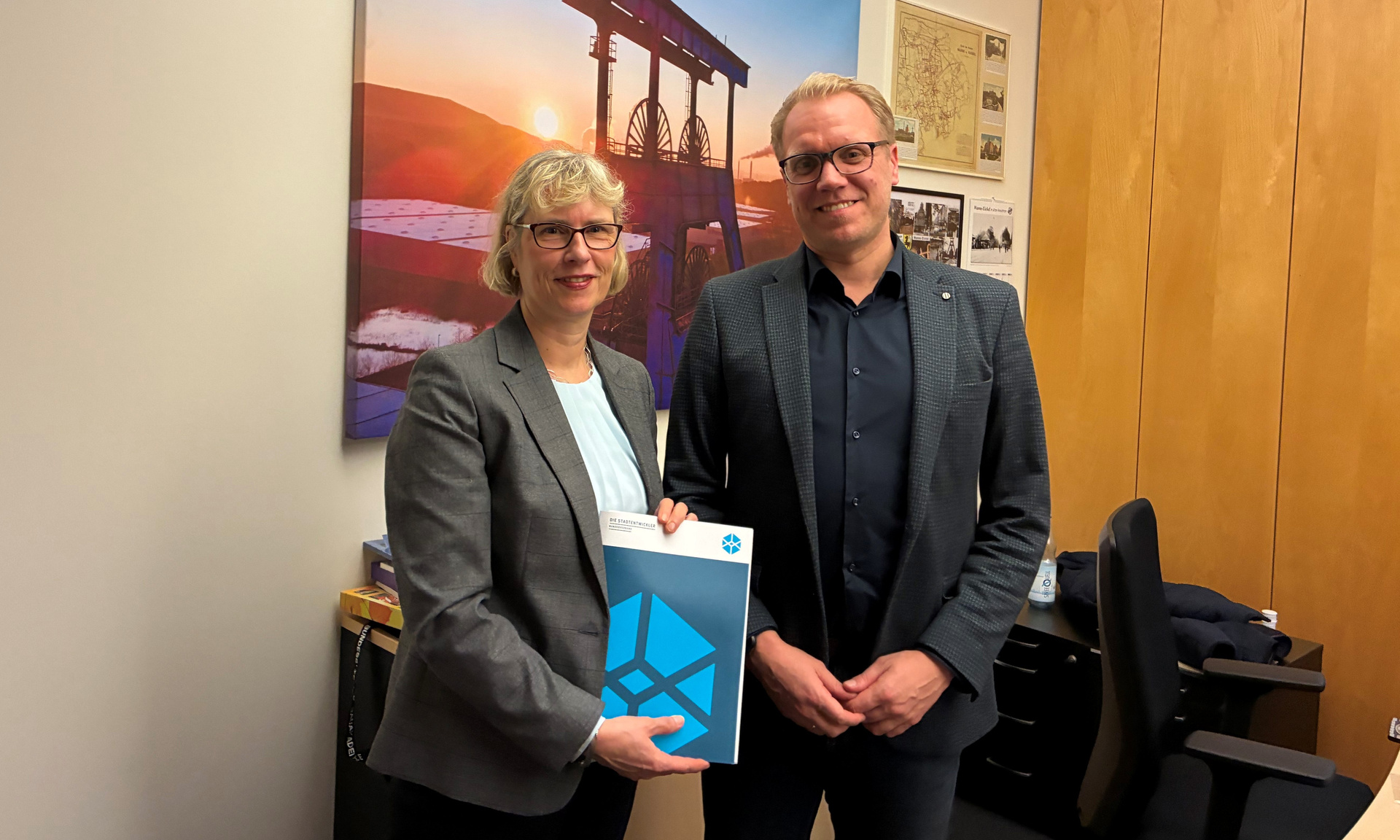 Monika Fontaine-Kretschmer und Hendrik Bollmann stehen mit dem Empfehlungspapier posierend der Kamera zugewandt.