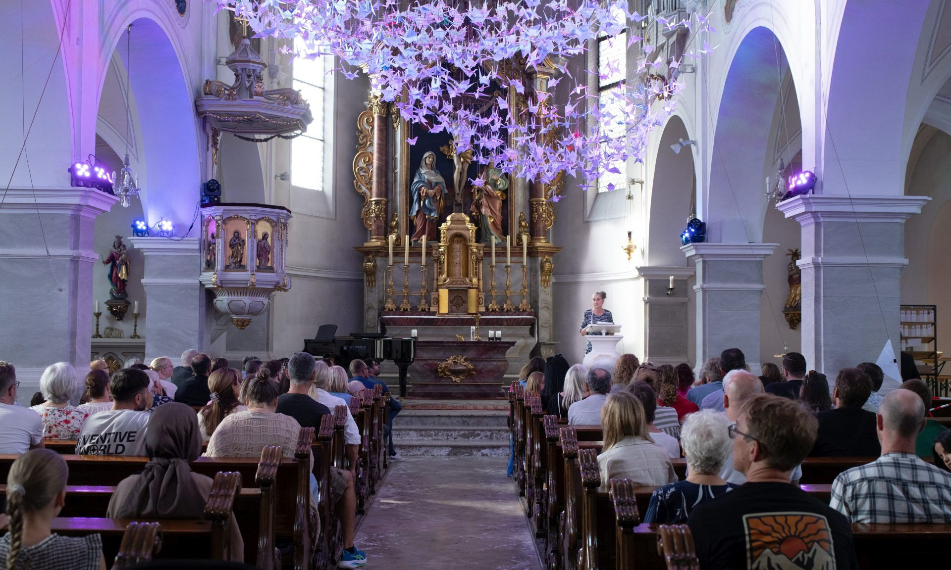 Blick von hinten in den Kirchenraum der Herz-Jesu-Kirche auf gut gefüllte Besucherbänke, den illuminierten Altar sowie die 843 Origami-Kraniche, die über den Menschen schweben