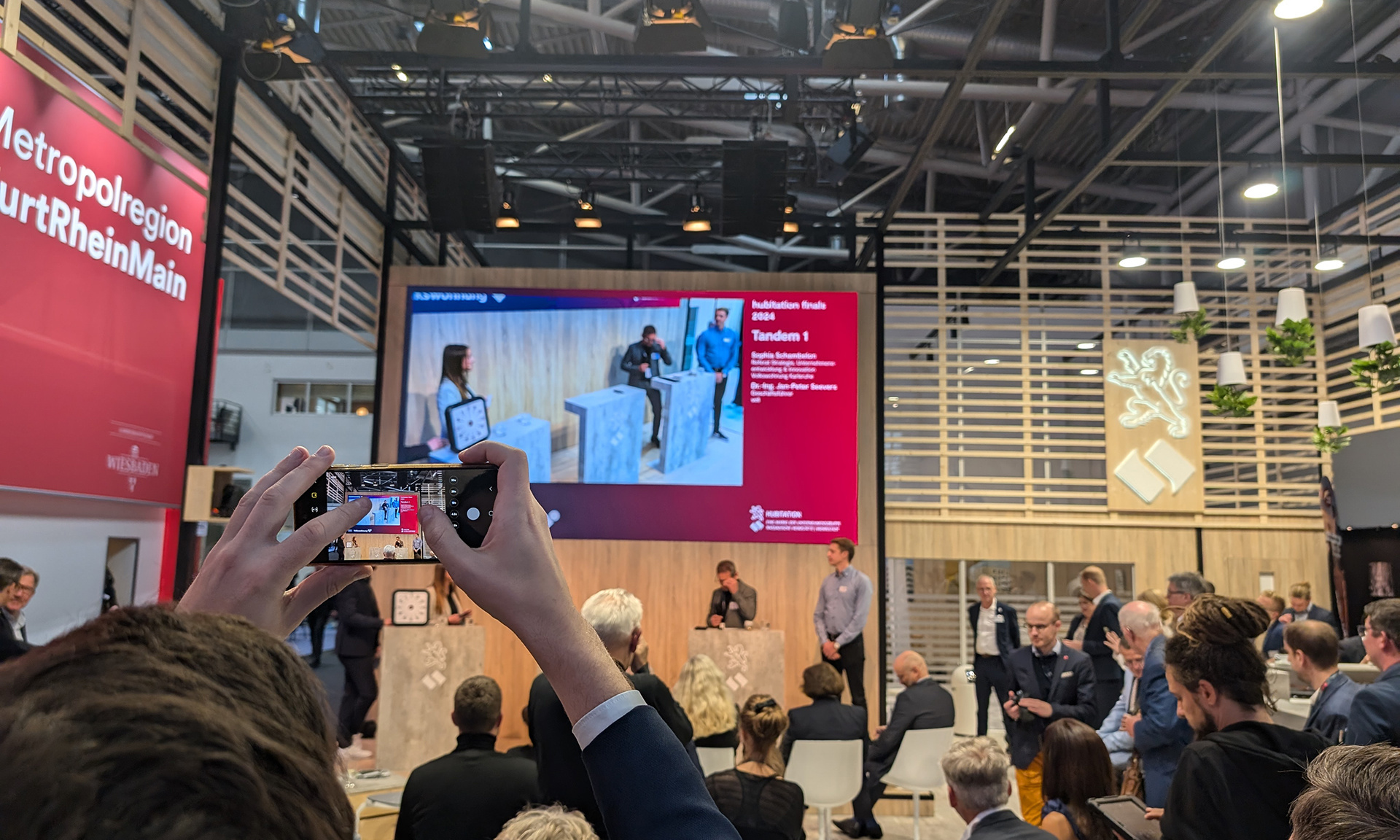 Zwei Hände halten ein Smartphone hoch, mit dem ein Schnappschuss von den hubitation finals gemacht wird. Jede Menge Publikum, im Hintergrund Wettbewerbsteilnehmer.