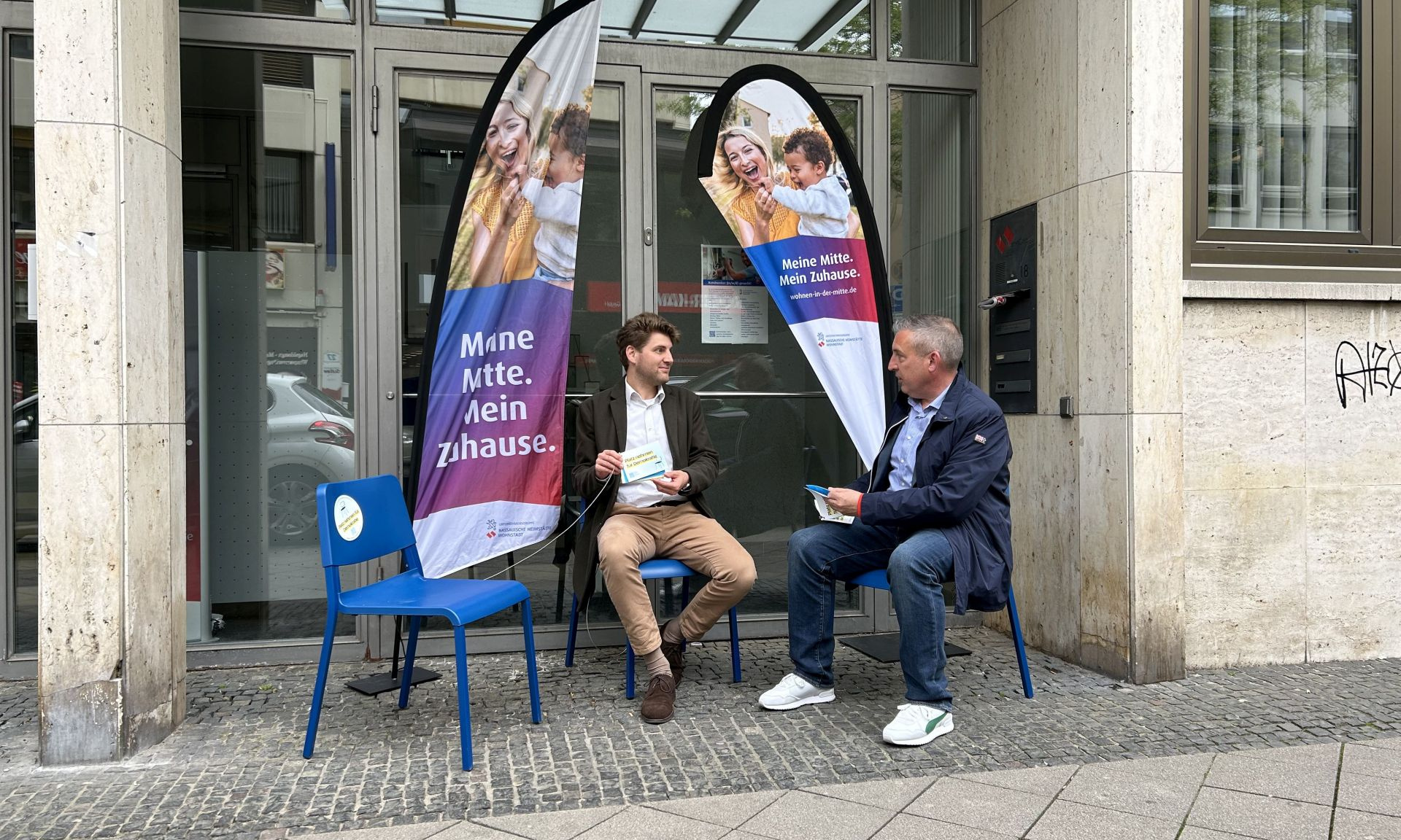 Zwei Männer sitzen auf Stühlen vor einem Bürogebäude und sprechen miteinander. 