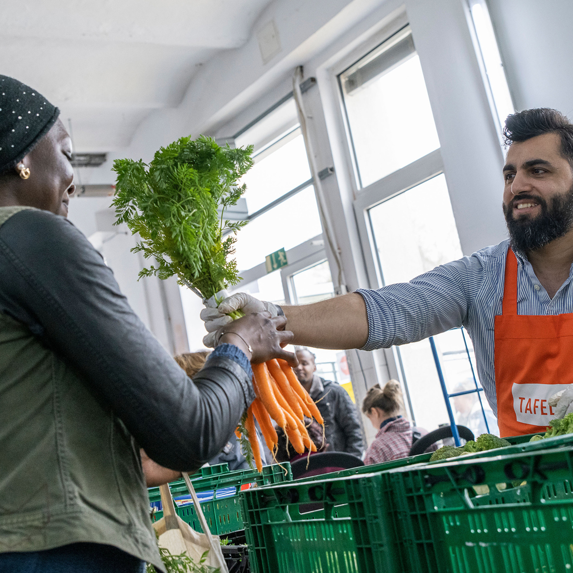 Übergabe bei einer Tafel. Mann gibt einer Frau ein Bund Karotten, vor ihm sind Kisten mit weiteren Lebensmitteln.