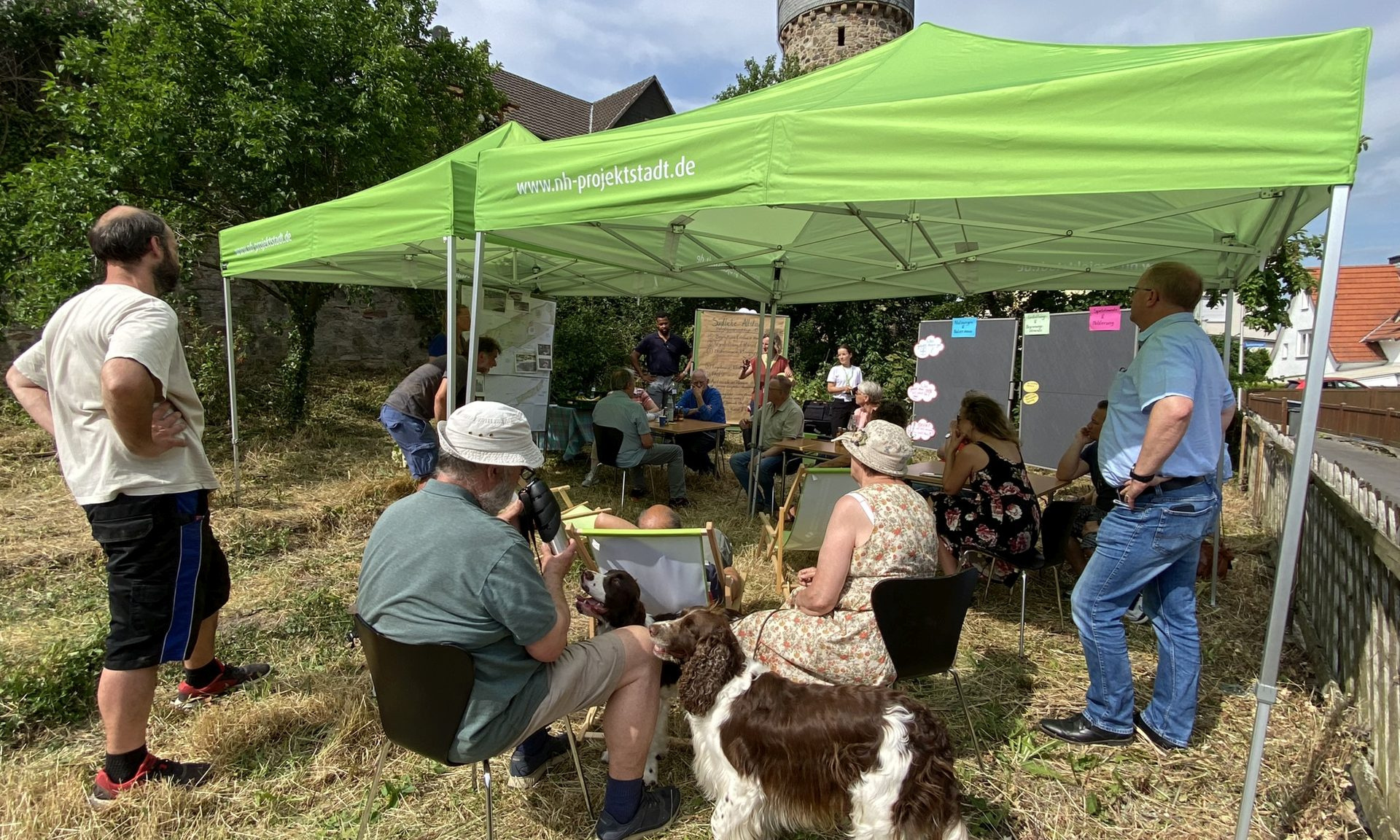 Eine Gruppe von Menschen sitzt unter einem Pavillon mit grünem Dach und der Aufschrift NH ProjektStadt