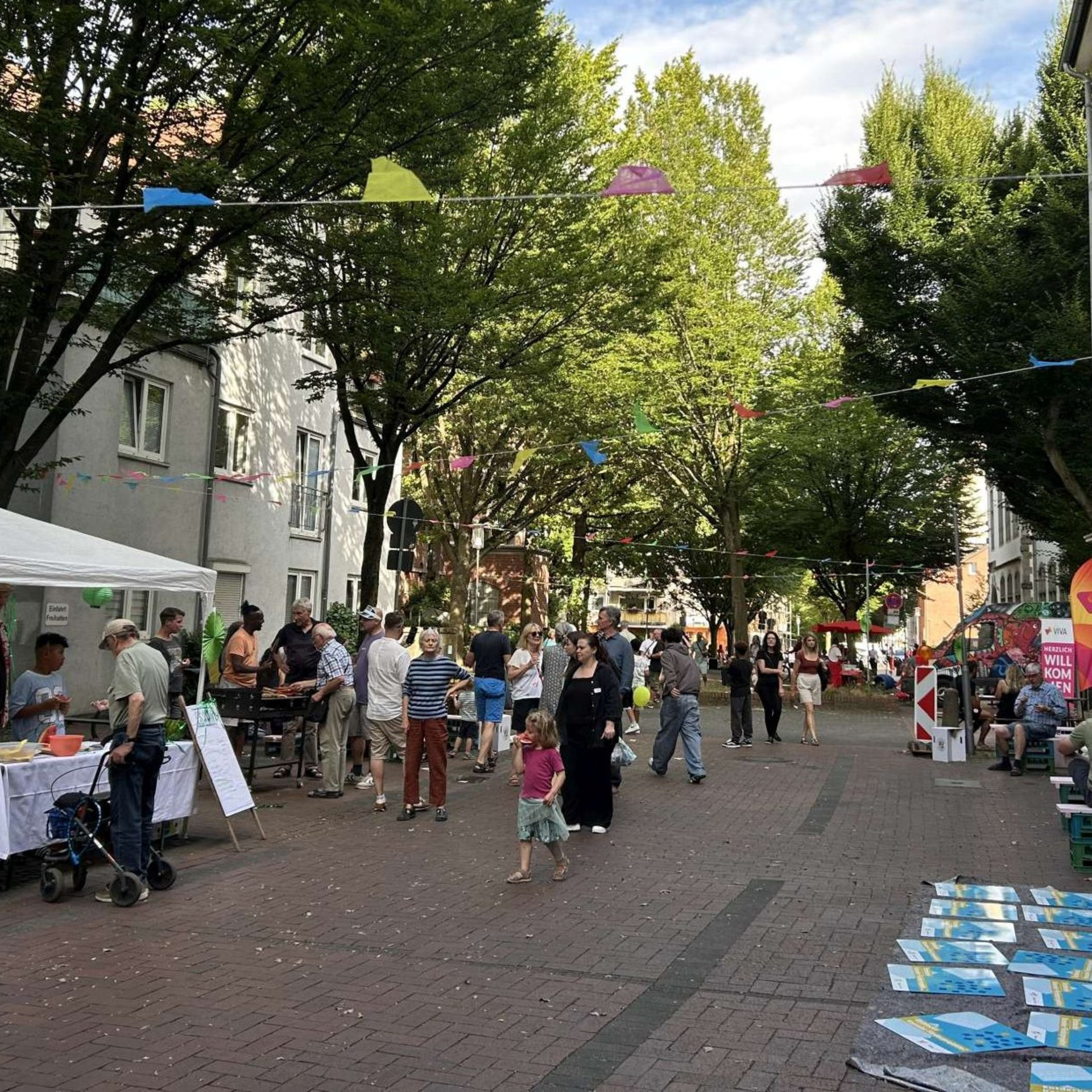 Mit Wimpeln geschmückte Straße, Menschen, die auf Bänken sitzen, vereinzelte Stände