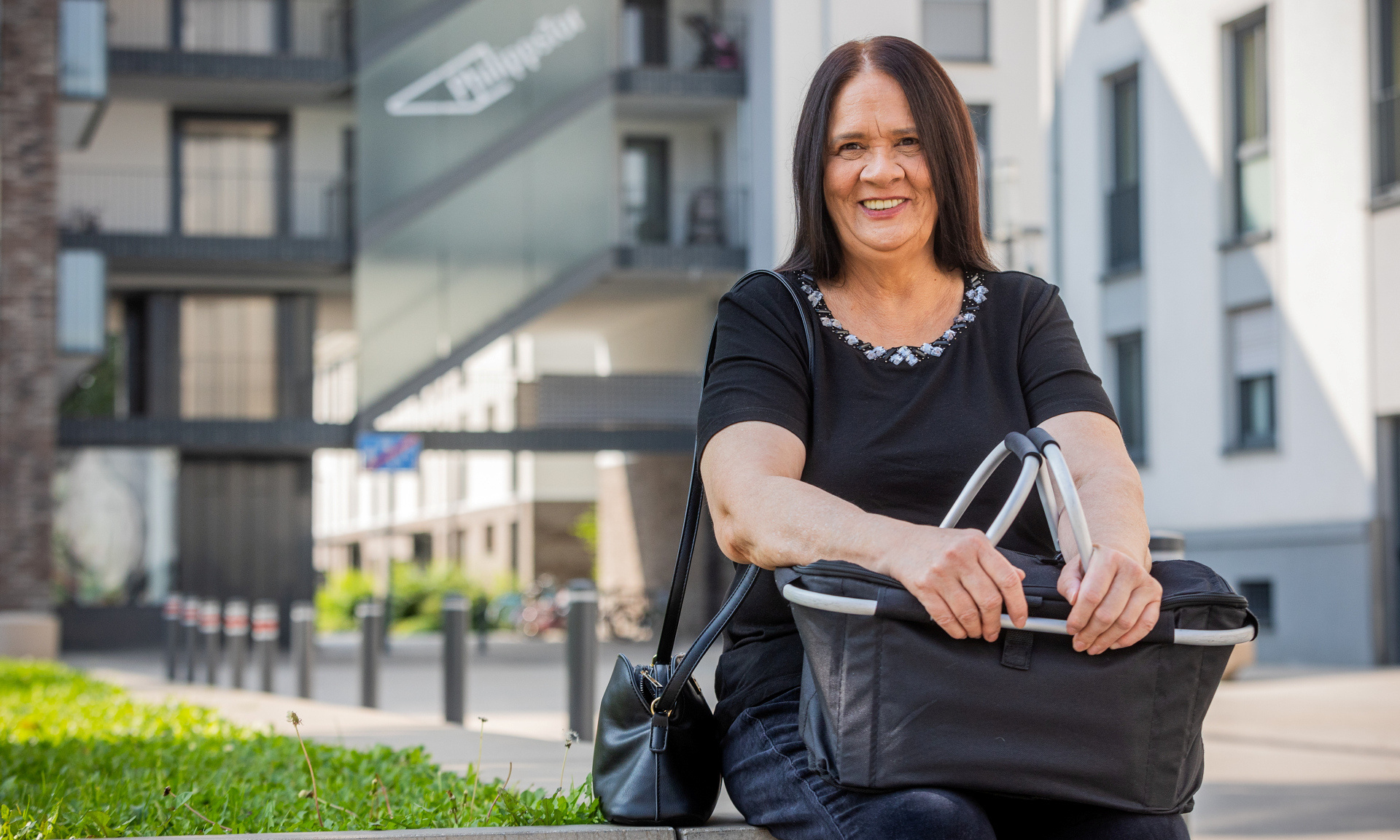 Mieterin sitzt vor einem Haus auf der Mauer und hat einen Einkaufskorb auf dem Schoß.