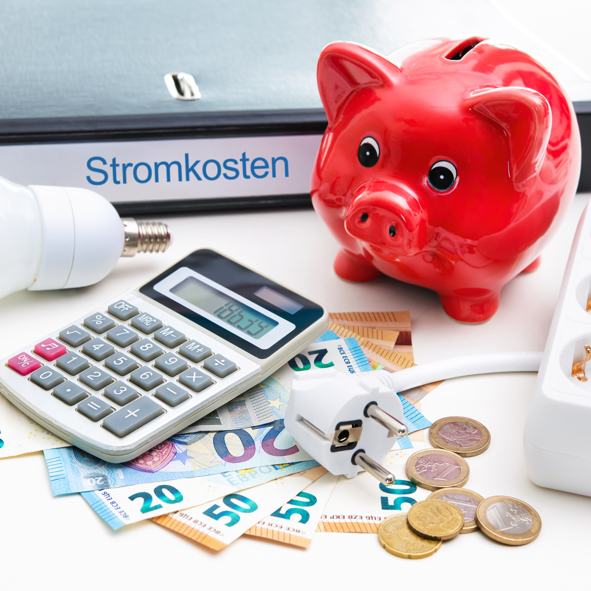 Auf einem Schreibtisch liegt Bargeld, ein Taschenrechner, ein Ordner auf dem Stromkosten steht und ein rotes Sparschwein