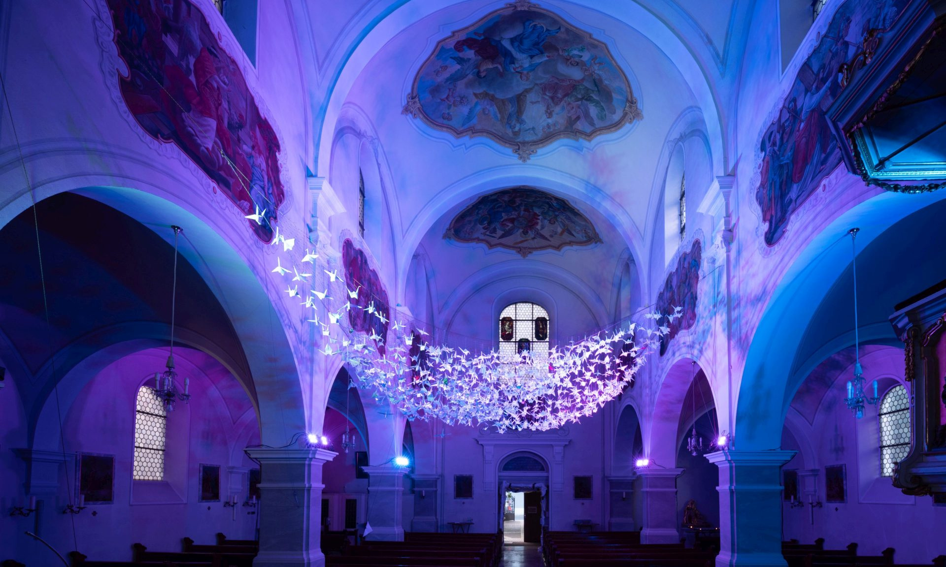 Blick in den leeren, lila illuminierten Kirchenraum der Herz-Jesu-Kirche in Kelsterbach mit den "schwebenden" 843 Origami-Kranichen