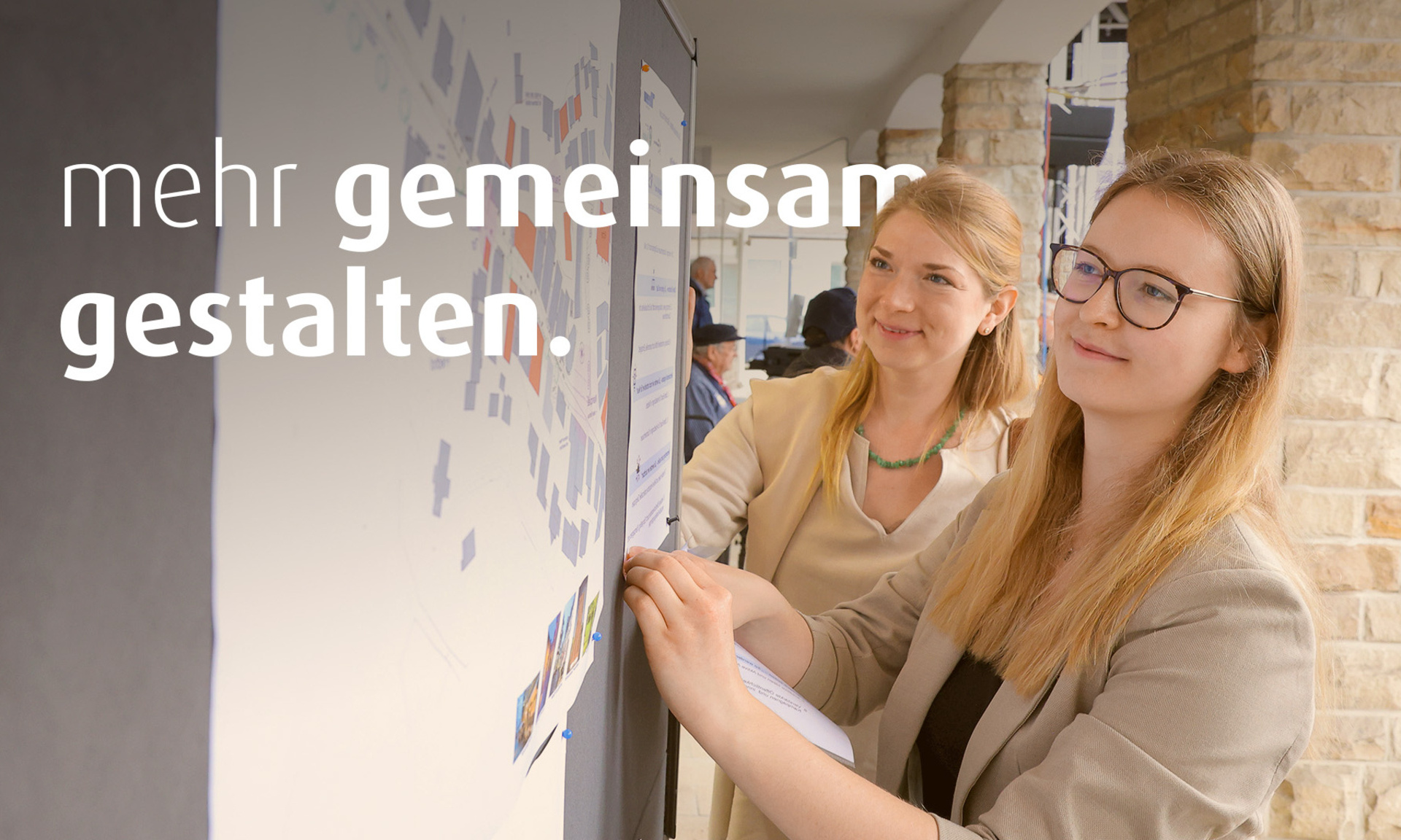 Zwei junge Frauen schauen auf einen Plan, der an der Wand befestigt ist. Auf dem Bild steht der Text "mehr gemeinsamer gestalten".