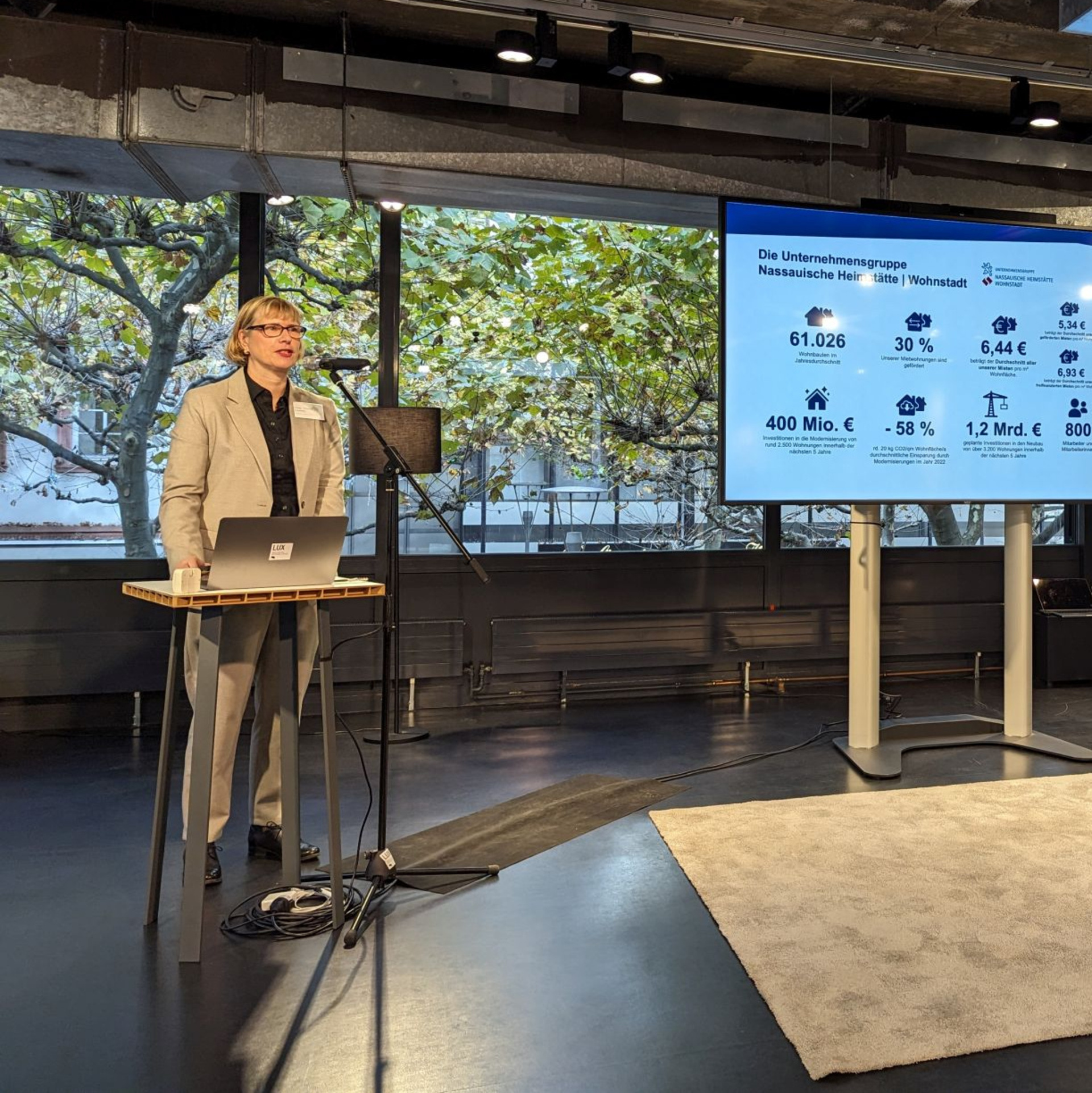 Monika Fontaine-Kretschmer spricht in ein Mikrofon, daneben ein Screen mit Infos zur NHW