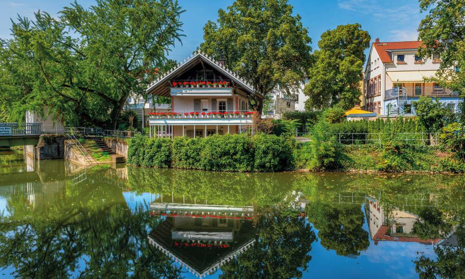 im Vordergrund See und weiter hinten ein kleines Haus mit Spitzdach und Terrassen