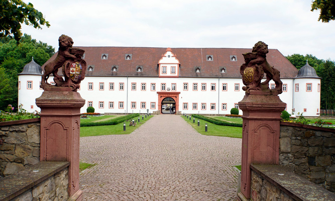 Blick auf ein schlossähnliches Gebäude mit einer gekiesten Auffahrt und zwei steinernen Figuren auf zwei Sockeln im Vordergrund