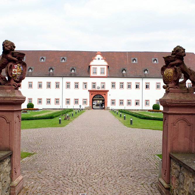 Blick auf ein schlossähnliches Gebäude mit einer gekiesten Auffahrt und zwei steinernen Figuren auf zwei Sockeln im Vordergrund