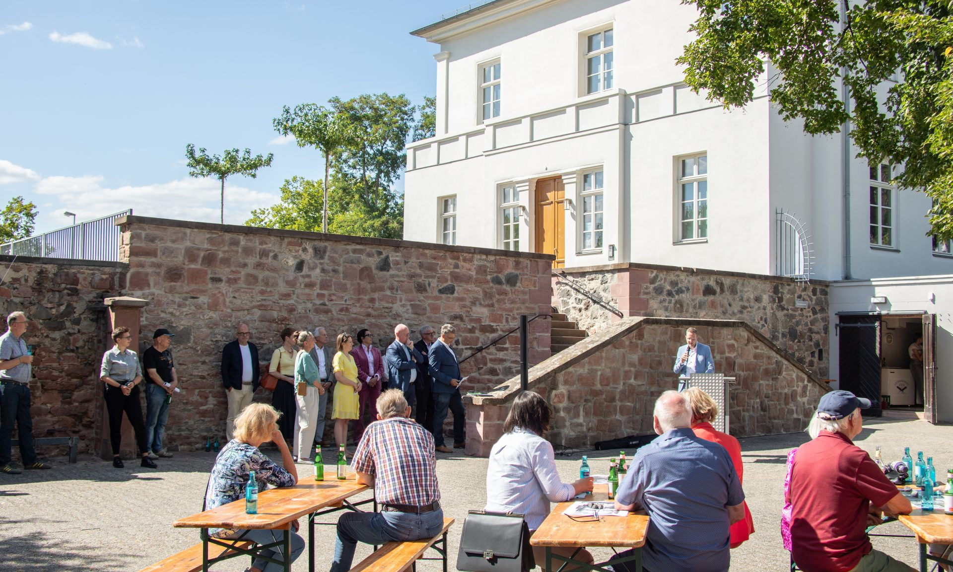 Menschen sitzen auf Bierbänken, im Hintergrund hält einer eine Ansprache, ganz hinten Schloss und Schlossmauer