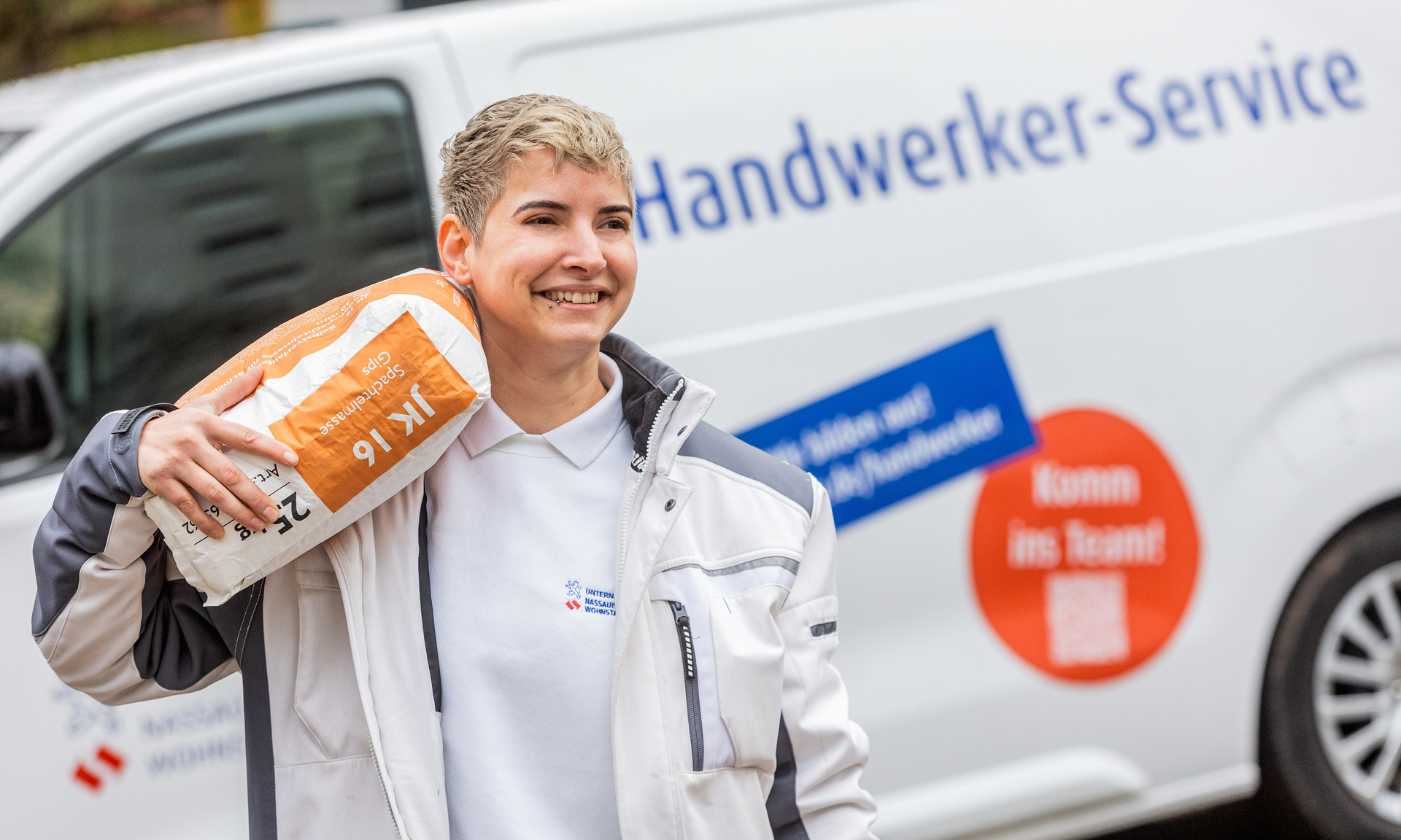 Das Bild zeigt eine Handwerkerin mit kurzen blonden Haaren, die lächelnd einen Sack auf der Schulter trägt, im Hintergrund ist ein Fahrzeug des NHW Handwerker Service zu sehen