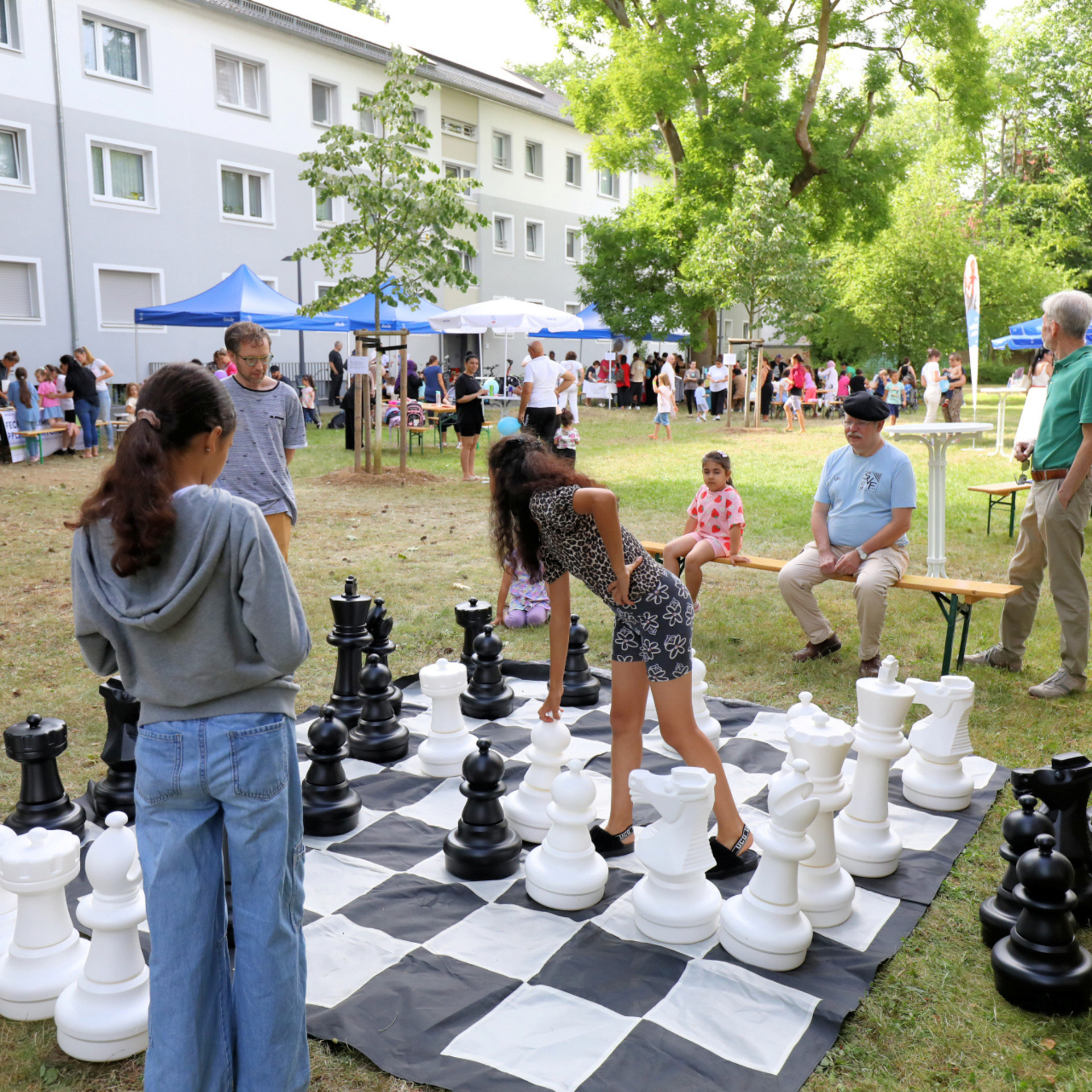 Kinder stehen um ein großes Schachbrett herum und spielen Schach. Im Hintergrund sind weitere Veranstaltungszelte zu sehen.
