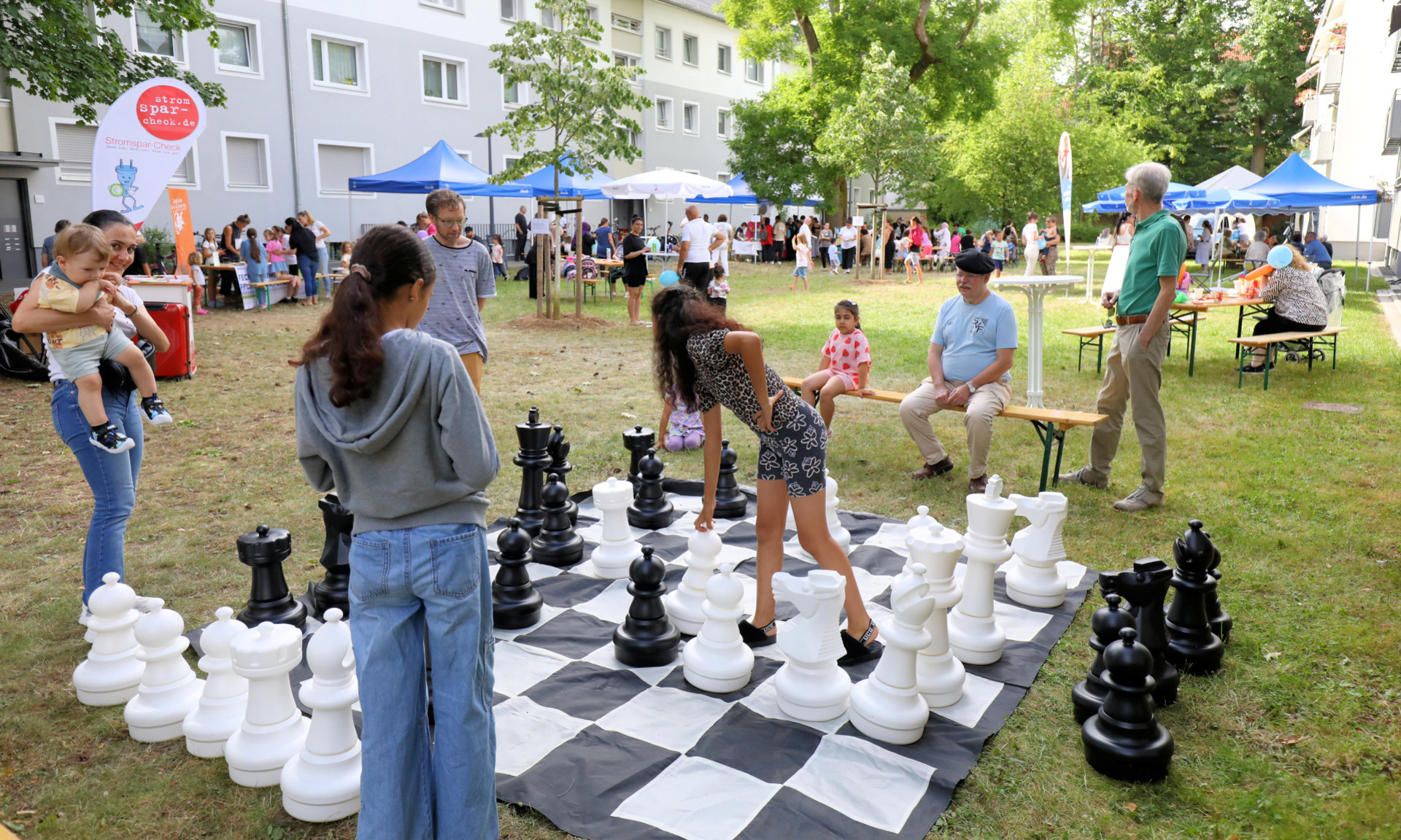 Kinder stehen um ein großes Schachbrett herum und spielen Schach. Im Hintergrund sind weitere Veranstaltungszelte zu sehen.