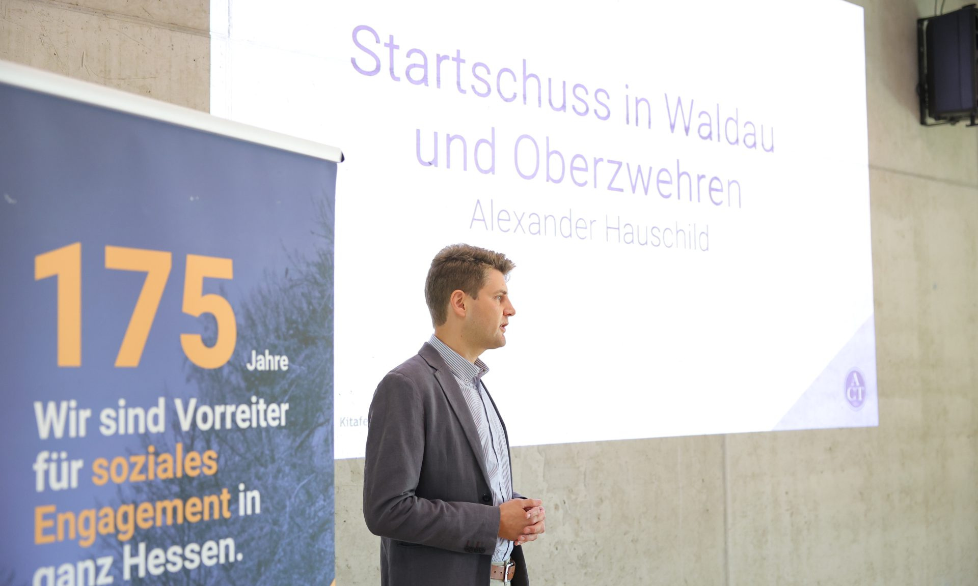 Alexander Hauschild (Soziale Quartiersentwicklung NHW) spricht, im Hintergrund ein Screen mit der Aufschrift "Startschuss in Waldau und Oberzwehren" sowie ein Banner mit der Aufschrift "175 Jahre. Wir sind Vorreiter für soziales Engagement in ganz Hessen."