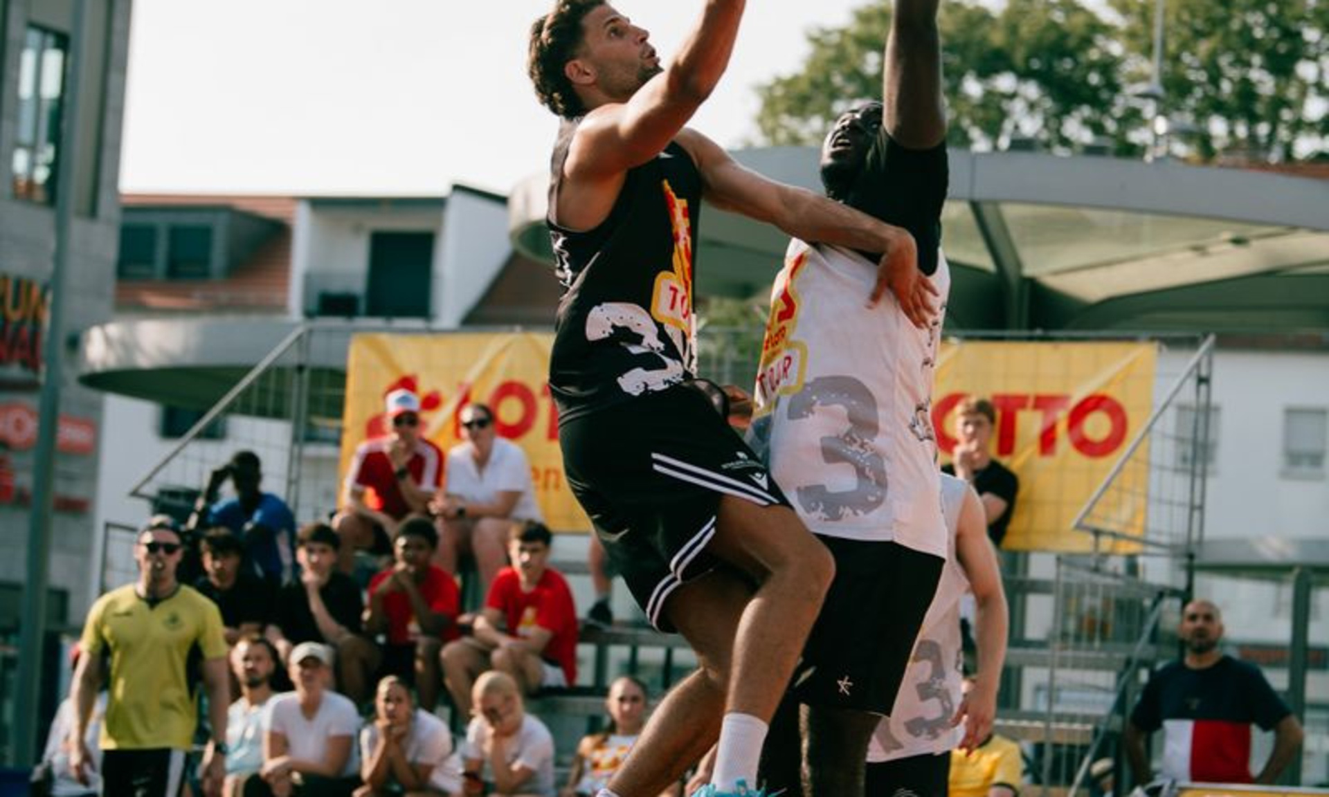 Zwei Streetbasketballspieler steigen zum Korb hoch, einer mit Ball, einer ohne, im Hintergrund Zuschauer auf einer Tribüne