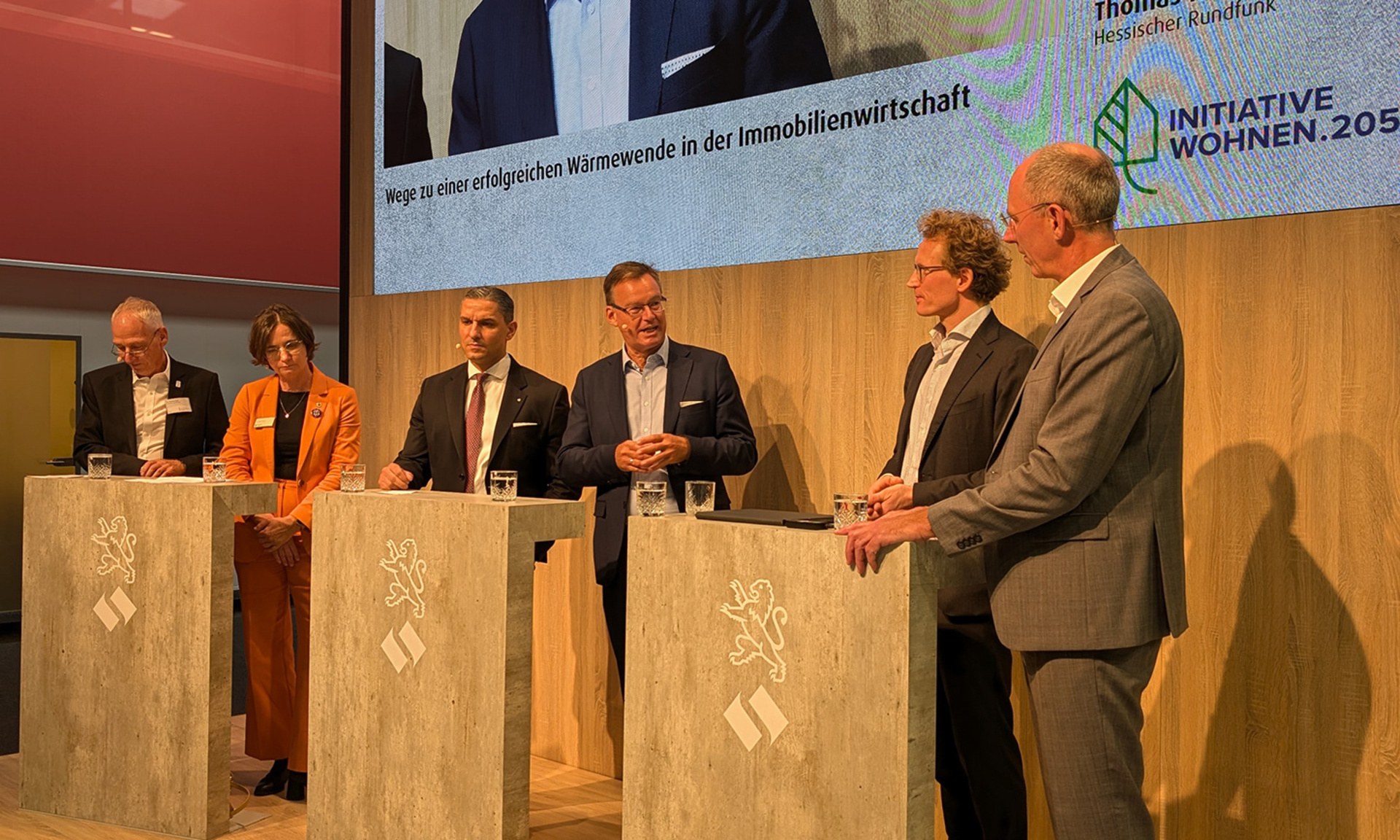 Sechs Menschen stehen hinter Rednerpulten und diskutieren miteinander, GdW-Präsident Axel Gedaschko wird im Screen darüber groß gezeigt