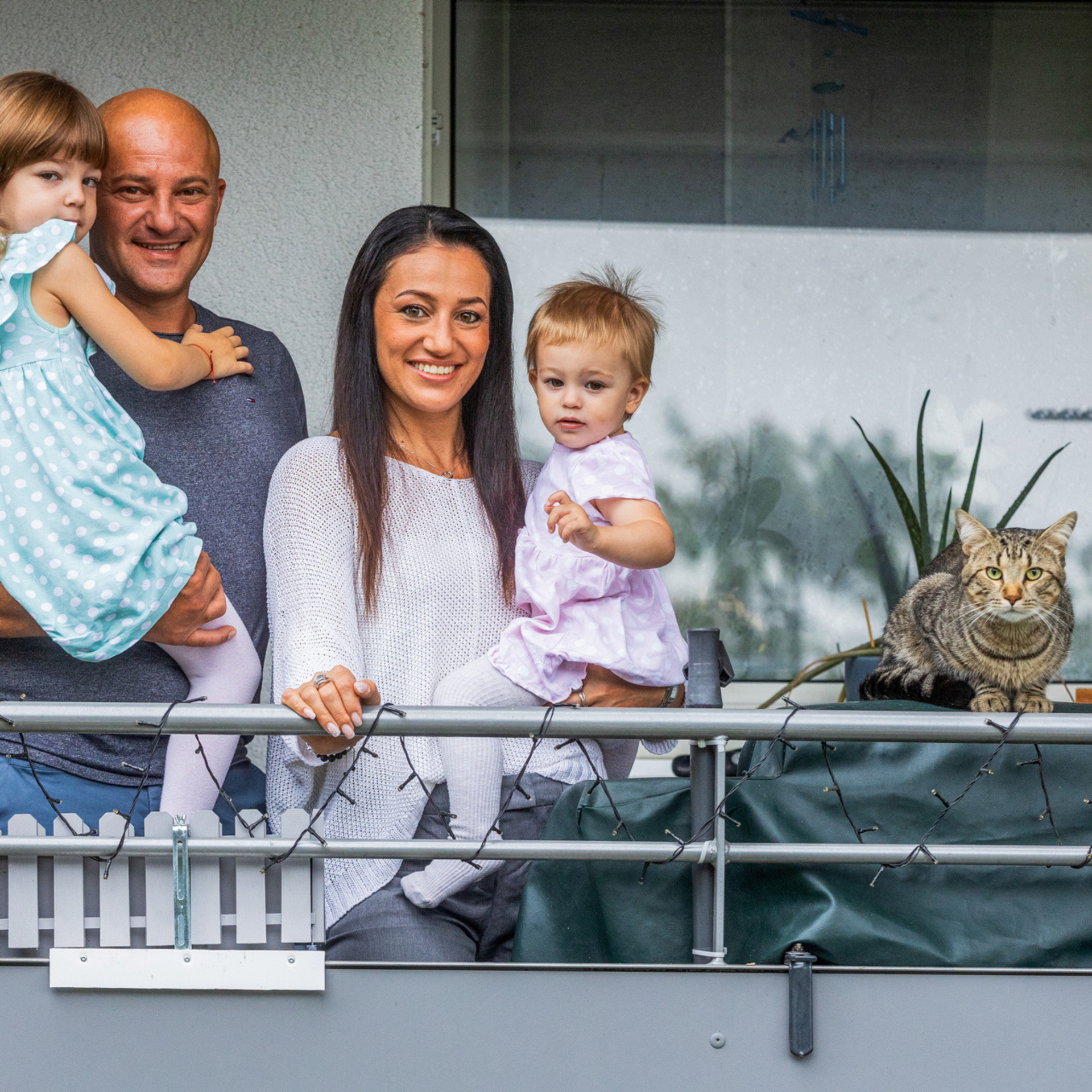 Familie steht an einem Balkon und schaut in die Kamera. Mann hält kleine Tochter im Arm, die Frau hält ein Baby im Arm.