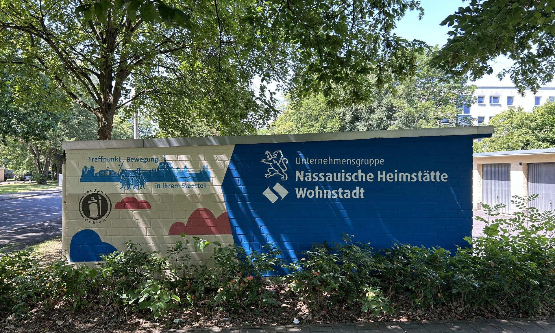 Eine bunt bemalte Wand, links mit dem Schriftzug "Treffpunkt Bewegung in Ihrem Stadtteil", rechts mit "Unternehmensgruppe Nassauische Heimstätte | Wohnstadt"