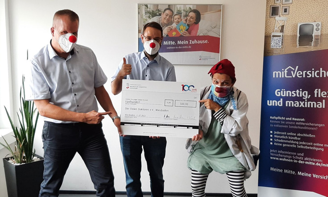 Zwei Männer übergeben einen Scheck an einen Clown