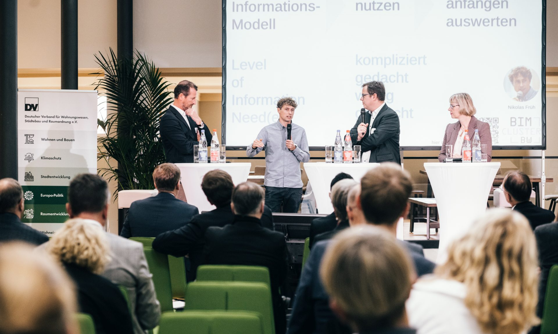 Blick auf eine Bühne mit vier Personen, zwei halten ein Mikrofon, davor Zuschauerreihen von hinten. Rechts NHW-Geschäftsführerin Monika Fontaine-Kretschmer