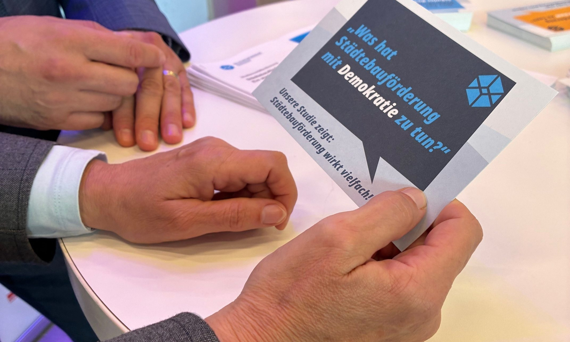 Eine Hand hält eine Karte mit einer eckigen Sprechblase und der Aufschrift "Was hat Städtebauförderung mit Demokratie zu tun?"