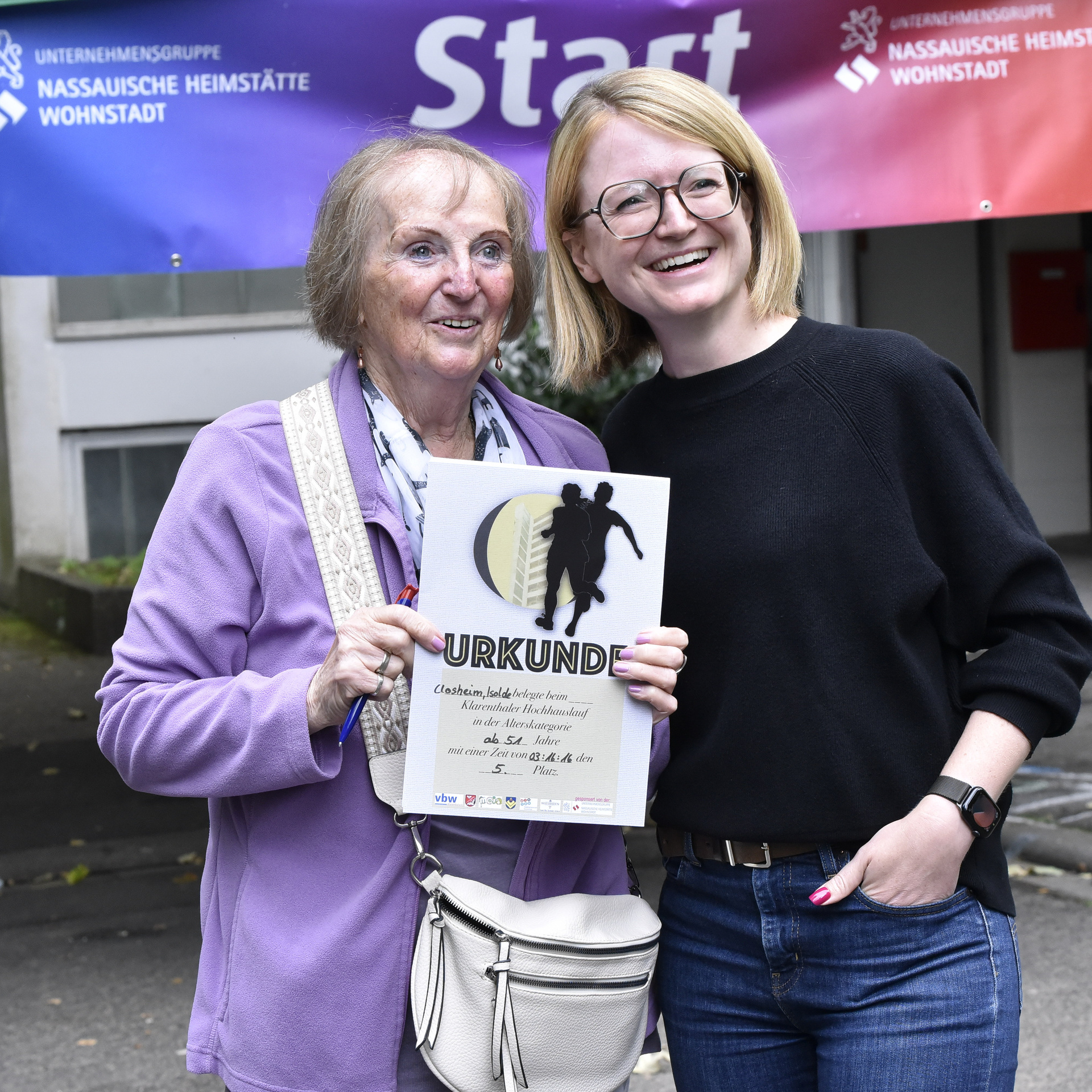 Seniorin, in sportlicher Kleidung, die eine Urkunde in der Hand hält und eine zweite Frau. Beide lächeln in die Kamera