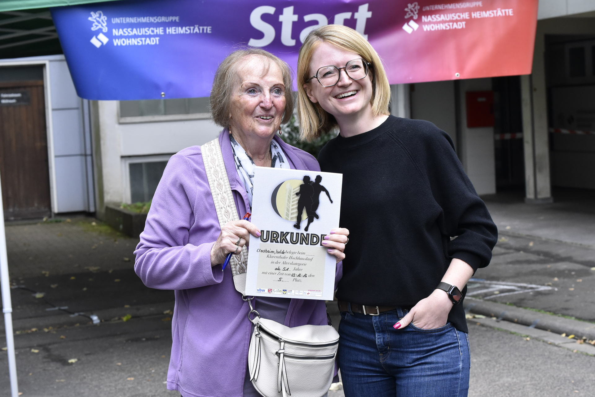 Seniorin, in sportlicher Kleidung, die eine Urkunde in der Hand hält und eine zweite Frau. Beide lächeln in die Kamera