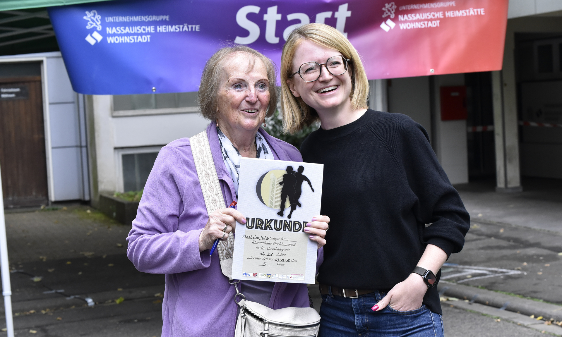 Seniorin, in sportlicher Kleidung, die eine Urkunde in der Hand hält und eine zweite Frau. Beide lächeln in die Kamera
