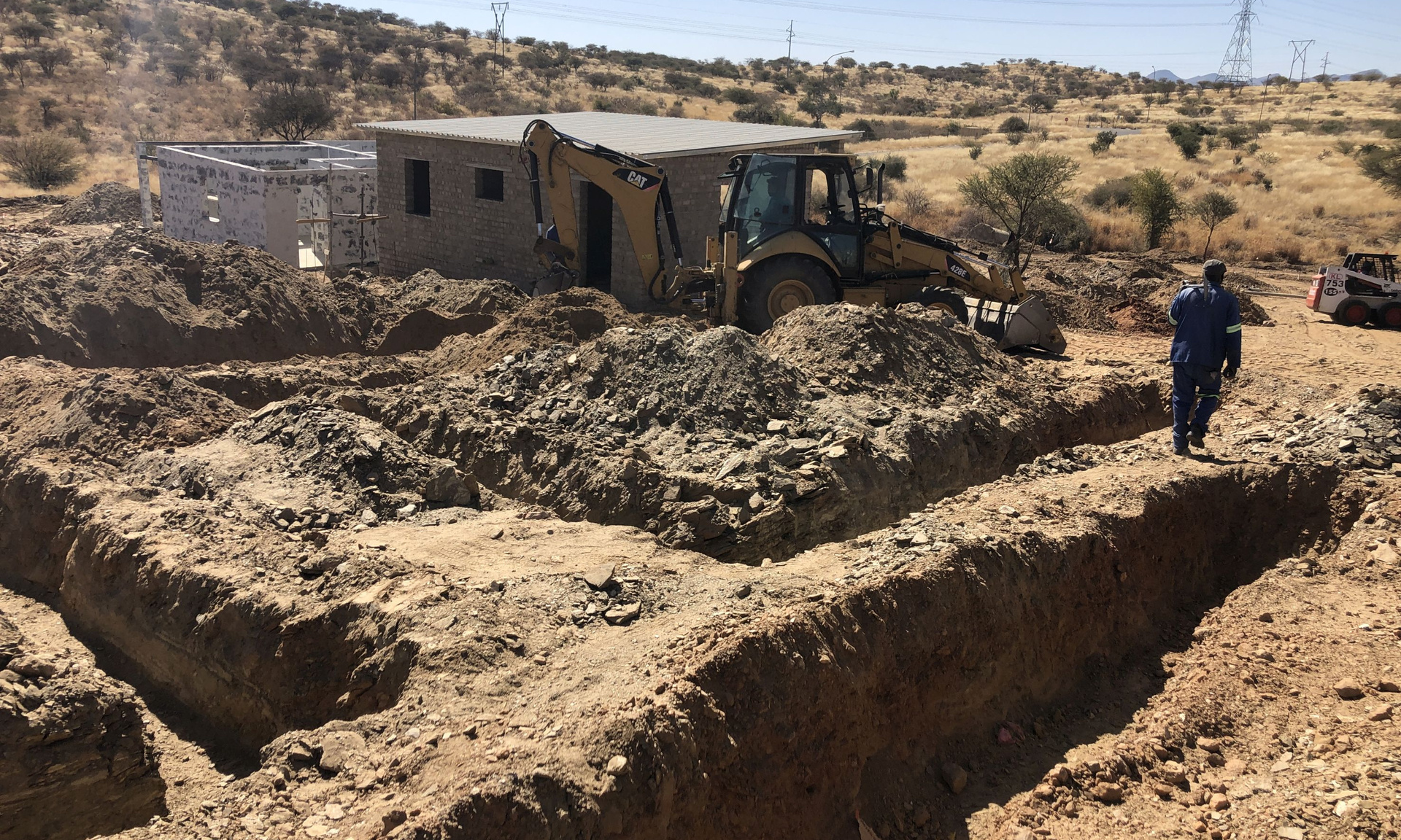 Ein Mann und ein Bagger auf einer Baustelle mit zwei Häusern, viel Sand und trockene Erde