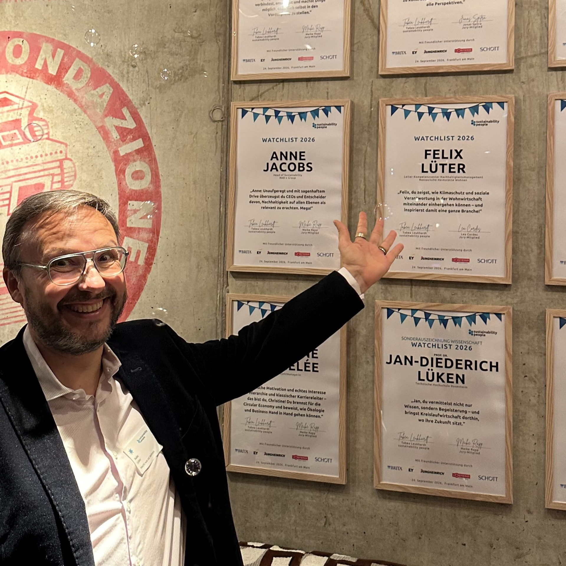 Felix Lüter, Leiter KC Nachhaltigkeitsmanagement bei der Unternehmensgruppe Nassauische Heimstätte | Wohnstadt, vor einer Wand mit Urkunden. Er zeigt auf die Urkunde mit seinem Namen.