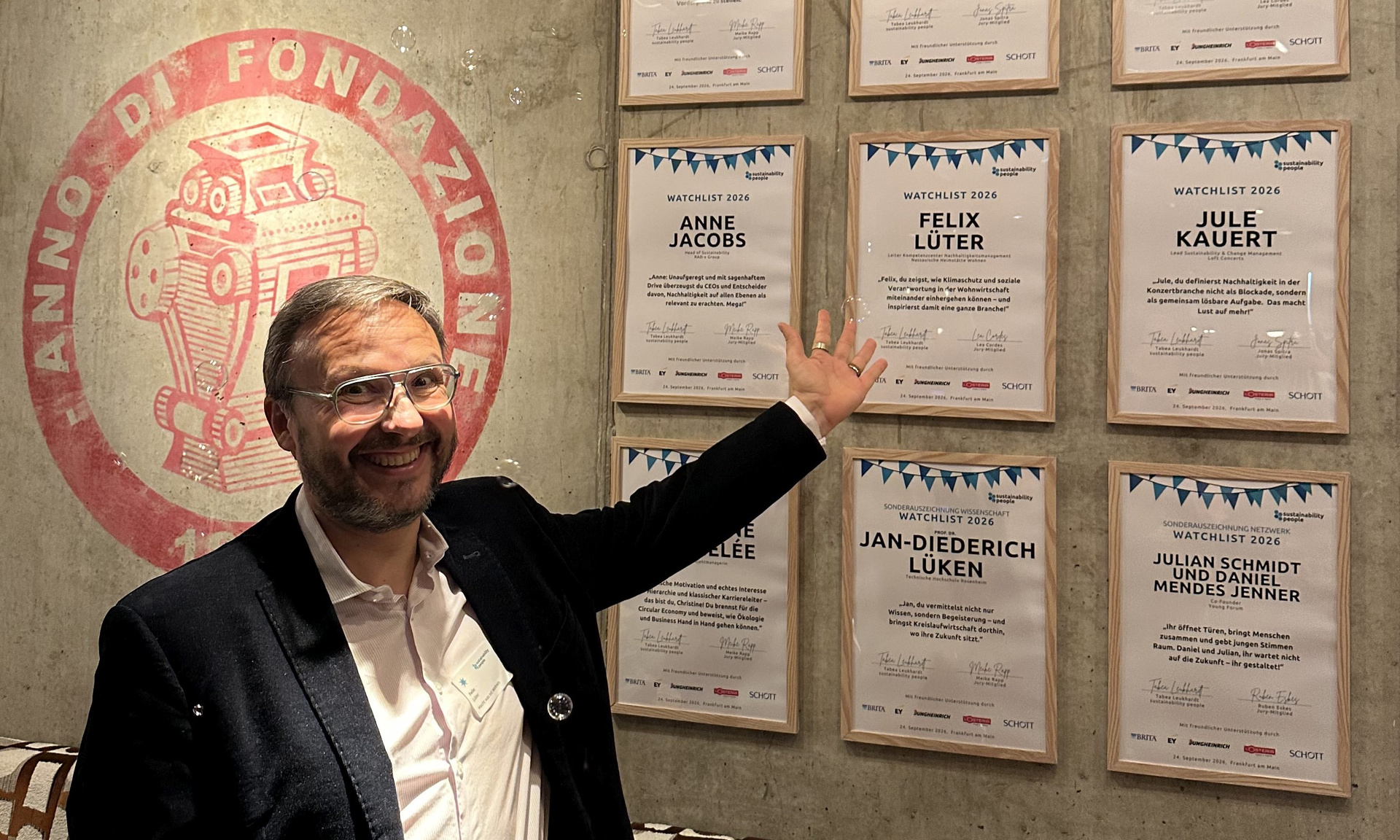 Felix Lüter, Leiter KC Nachhaltigkeitsmanagement bei der Unternehmensgruppe Nassauische Heimstätte | Wohnstadt, vor einer Wand mit Urkunden. Er zeigt auf die Urkunde mit seinem Namen.