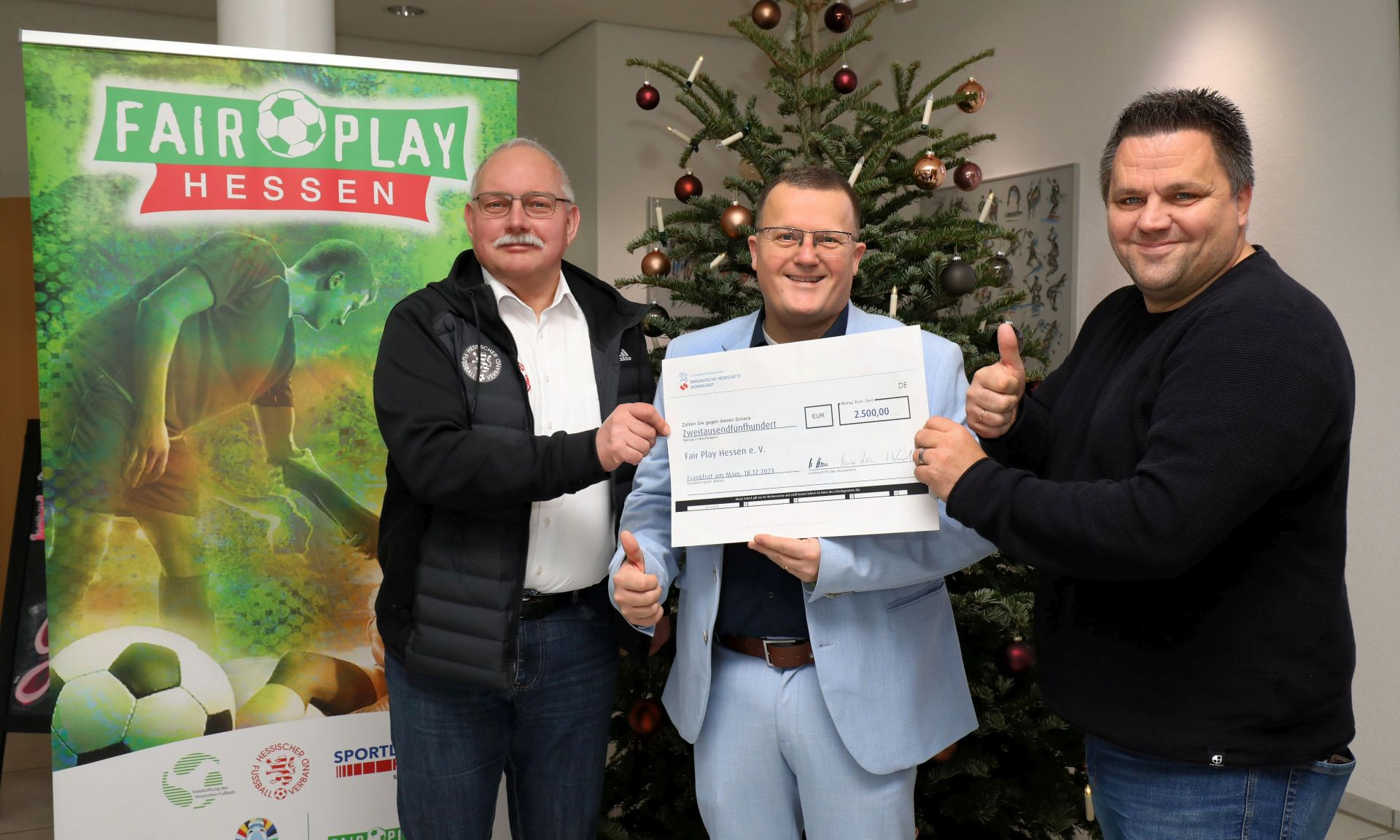 Drei Personen mit großem Spendenscheck vor einem Plakat mit der Aufschrift "Fairplay Hessen" sowie einem Weihnachtsbaum: Holger Lack (Mi., Regionalcenterleiter Frankfurt), übergibt die Weihnachtsspende der NHW für Südhessen an Thorsten Schenk (re.), Geschäftsführer der Sozialstiftung des Hessischen Fußballs, und Claus Menke (stellvertretender Vorsitzender der Sozialstiftung des Hessischen Fußballs sowie Vorsitzender des Verbandsausschusses für Freizeit- und Breitensport des Hessischen Fußball-Verbandes).
