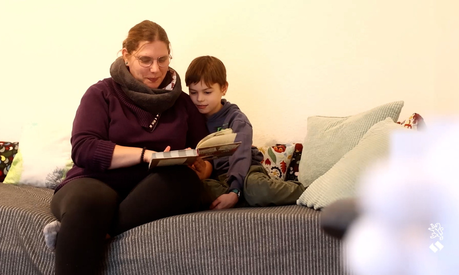 Eine Frau sitzt mit einem Jungen auf einem Bett. Gemeinsam schauen sie in ein Buch.