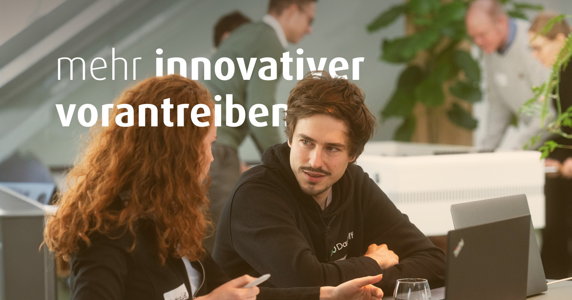 Eine Frau und ein Mann sitzen nebeneinander an einem Tisch. Vor ihnen stehen Laptops. Im Bild steht der Text "mehr innovativer vorantreiben".