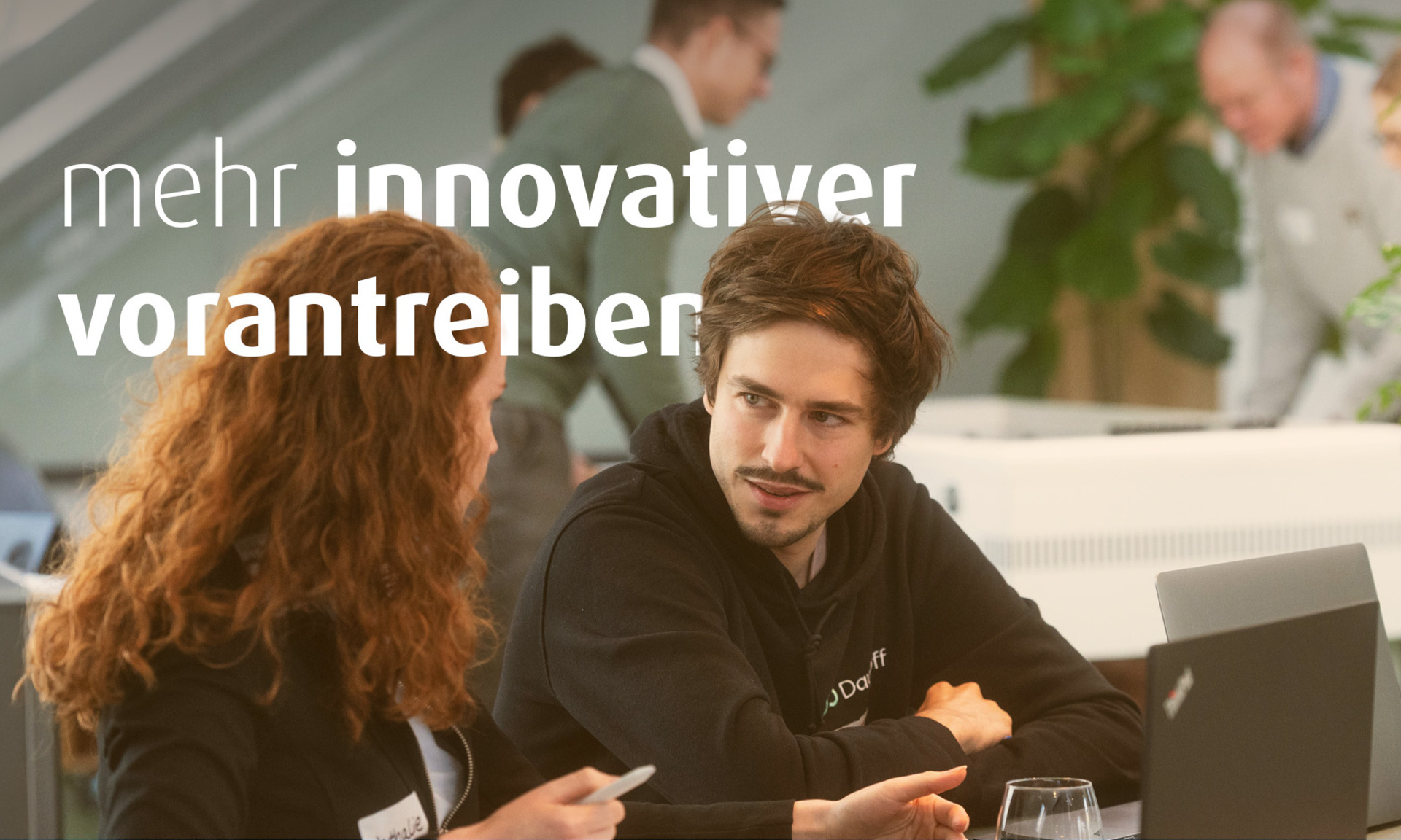 Eine Frau und ein Mann sitzen nebeneinander an einem Tisch. Vor ihnen stehen Laptops. Im Bild steht der Text "mehr innovativer vorantreiben".