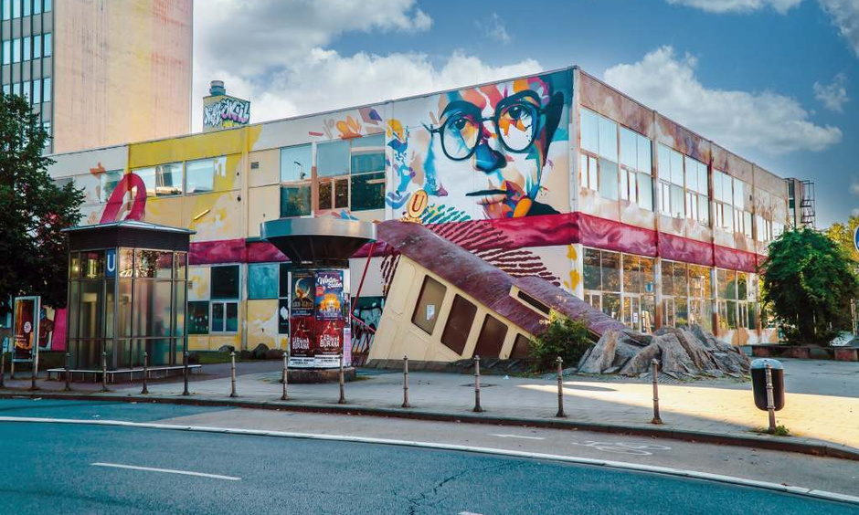 Gebäude mit bemalter Fassade, im Vordergrund U-Bahn Station, Zugang ist ein Bahnwagen der in die Erde geht