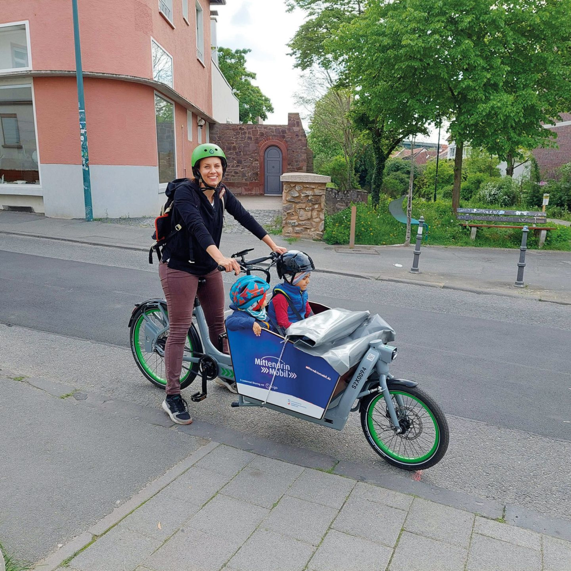 Frau mit zwei Kindern in einem Lastenrad. Alle tragen Helme.