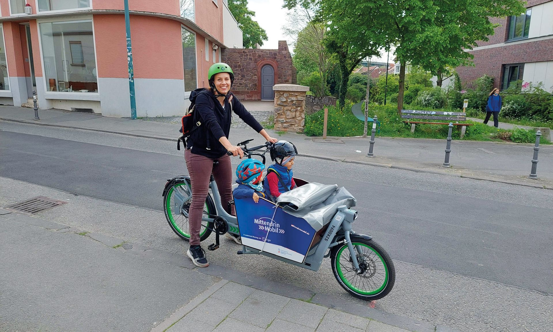 Frau mit zwei Kindern in einem Lastenrad. Alle tragen Helme.