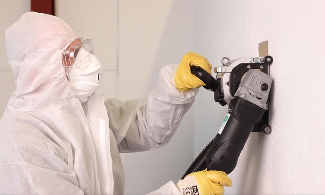 Mann in Schutzanzug entfernt maschinell Asbest von der Wand.