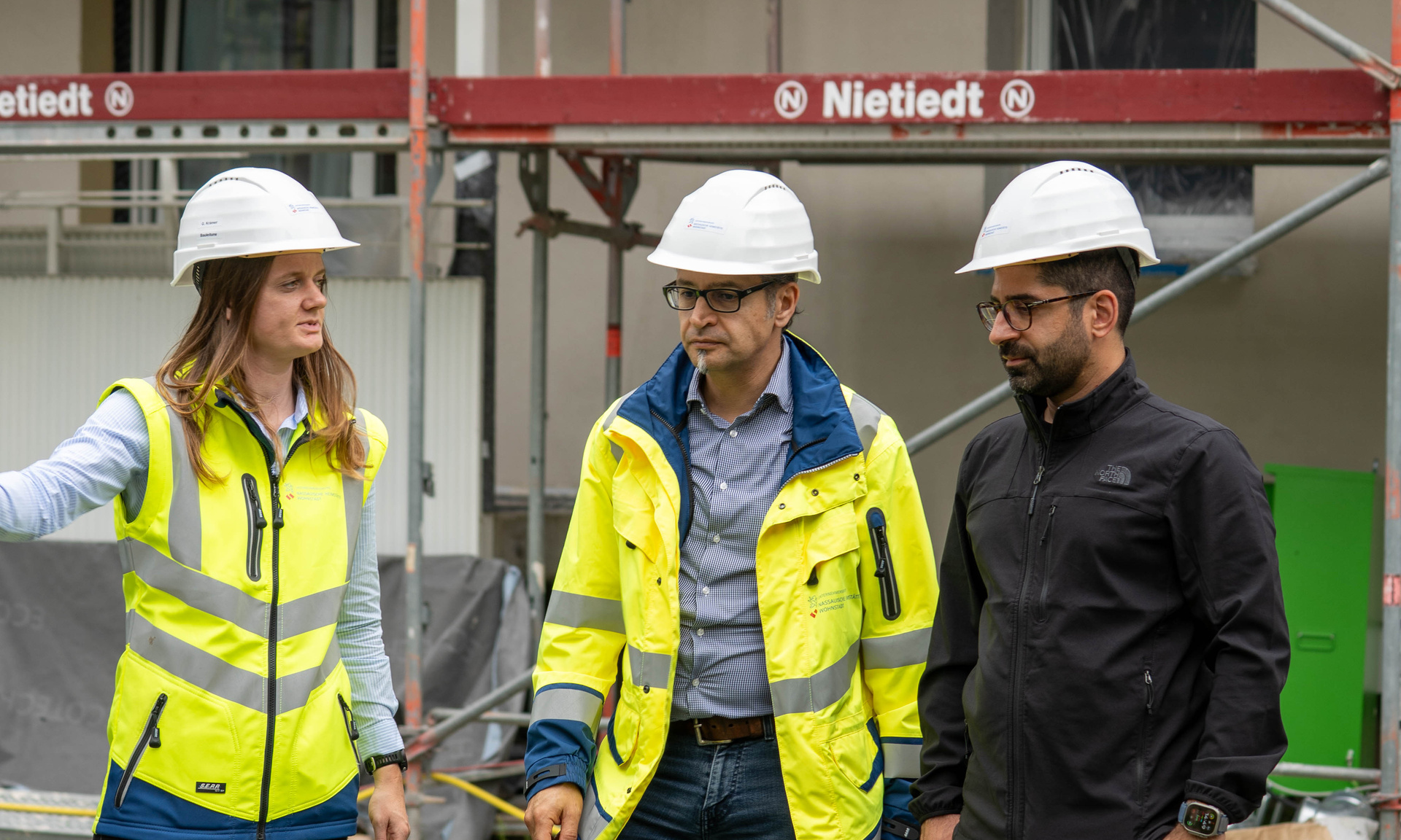 Frau und Mann mit Warnweste und weißen Bauhelm an. Dritte Person ohne Warnweste, schwarze Kleidung mit weißen Helm.