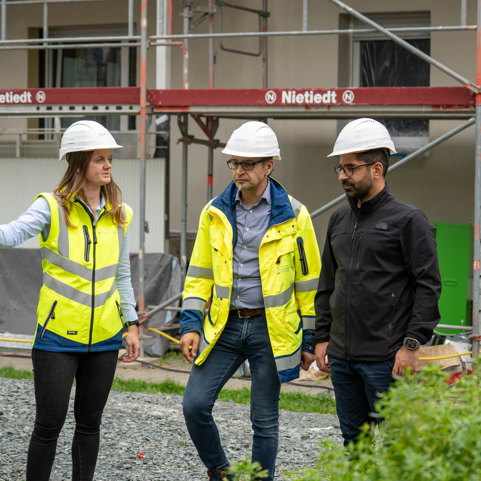 Frau und Mann mit Warnweste und weißen Bauhelm an. Dritte Person ohne Warnweste, schwarze Kleidung mit weißen Helm.