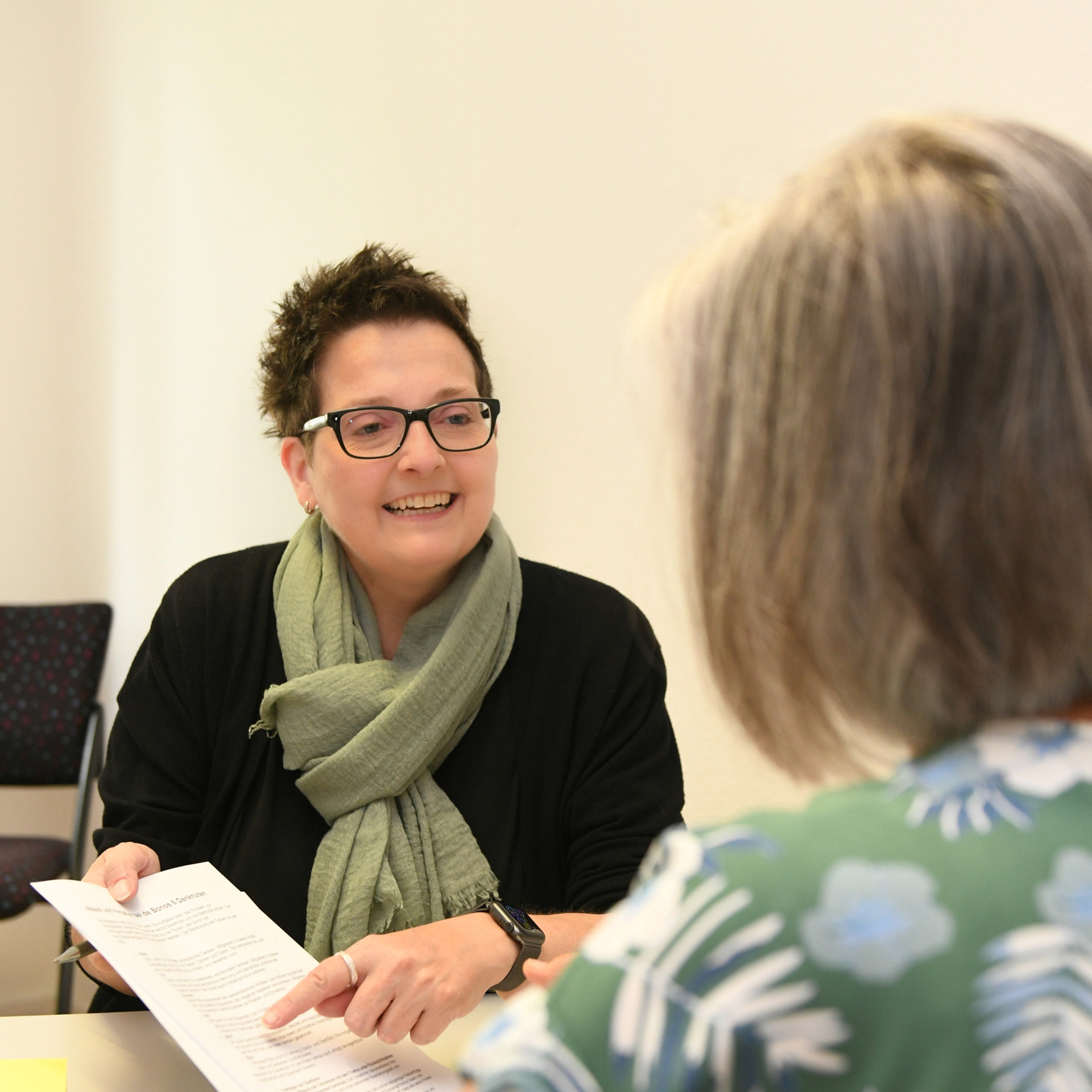 Frau Sitz an einem Schreibtisch mit einem Blatt in der Hand und berät eine andere Frau, die mit dem Rücken zur Kamera sitzt
